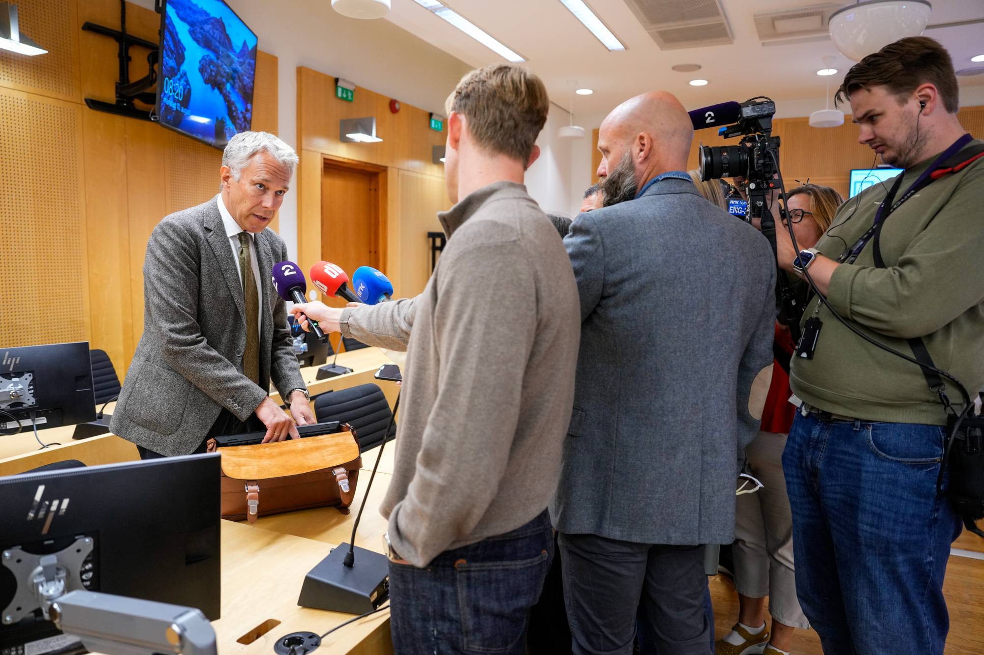 Mann i dress blir intervjuet av journalister med mikrofoner og kamera i et møterom. (Bildebeskrivelsen er laget av en KI-tjeneste)