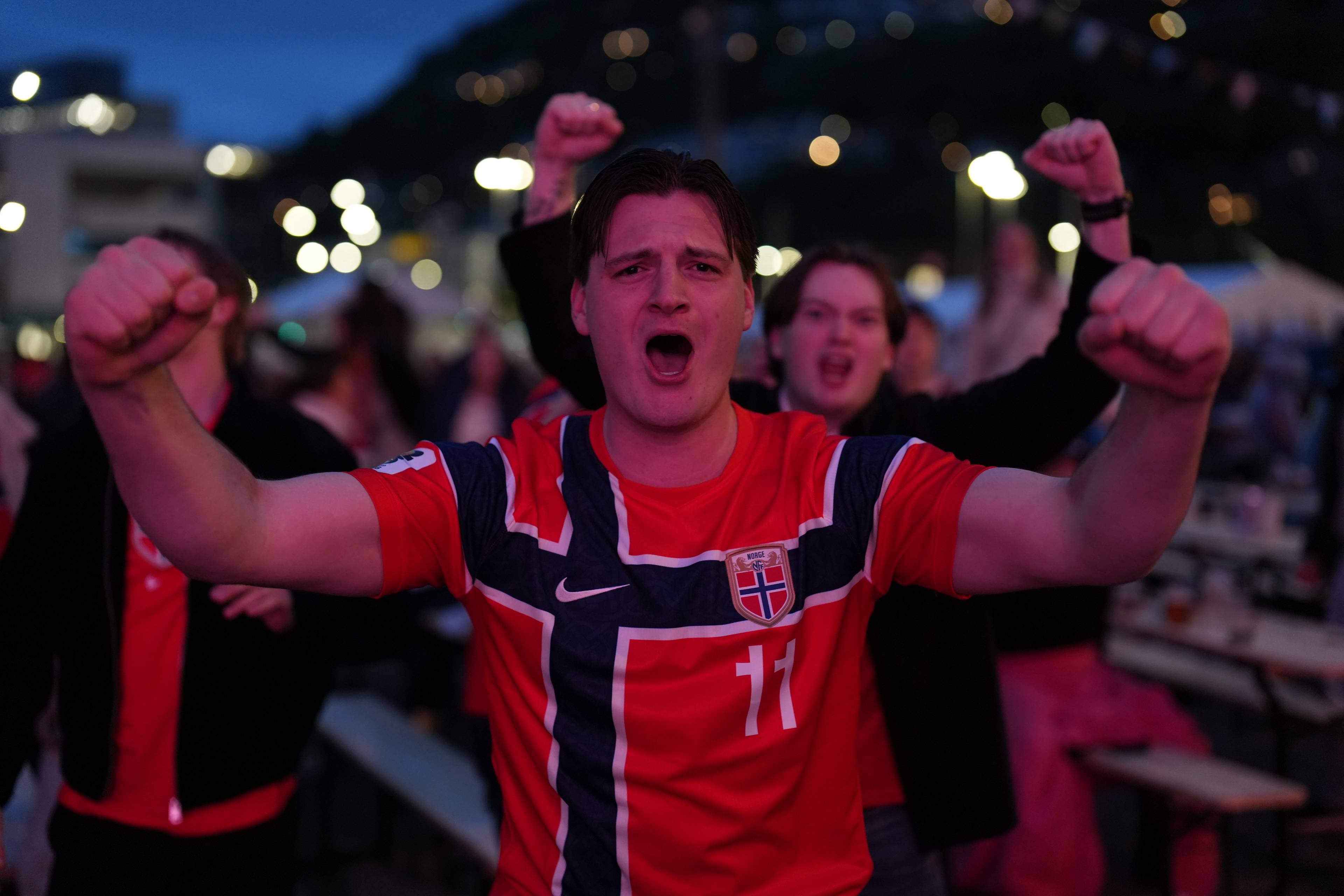 En mann i en norsk fotballtrøye jubler med knyttede never. Han er omringet av andre supporters i kveldslys. Flere personer kan ses i bakgrunnen.