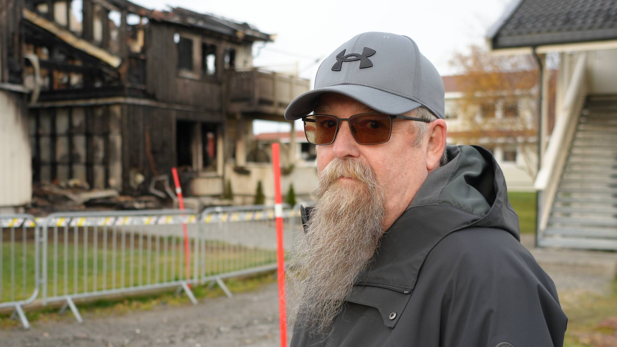 Mann med skjegg og caps står foran et brannskadet bygg avsperret med metallgjerder. 
