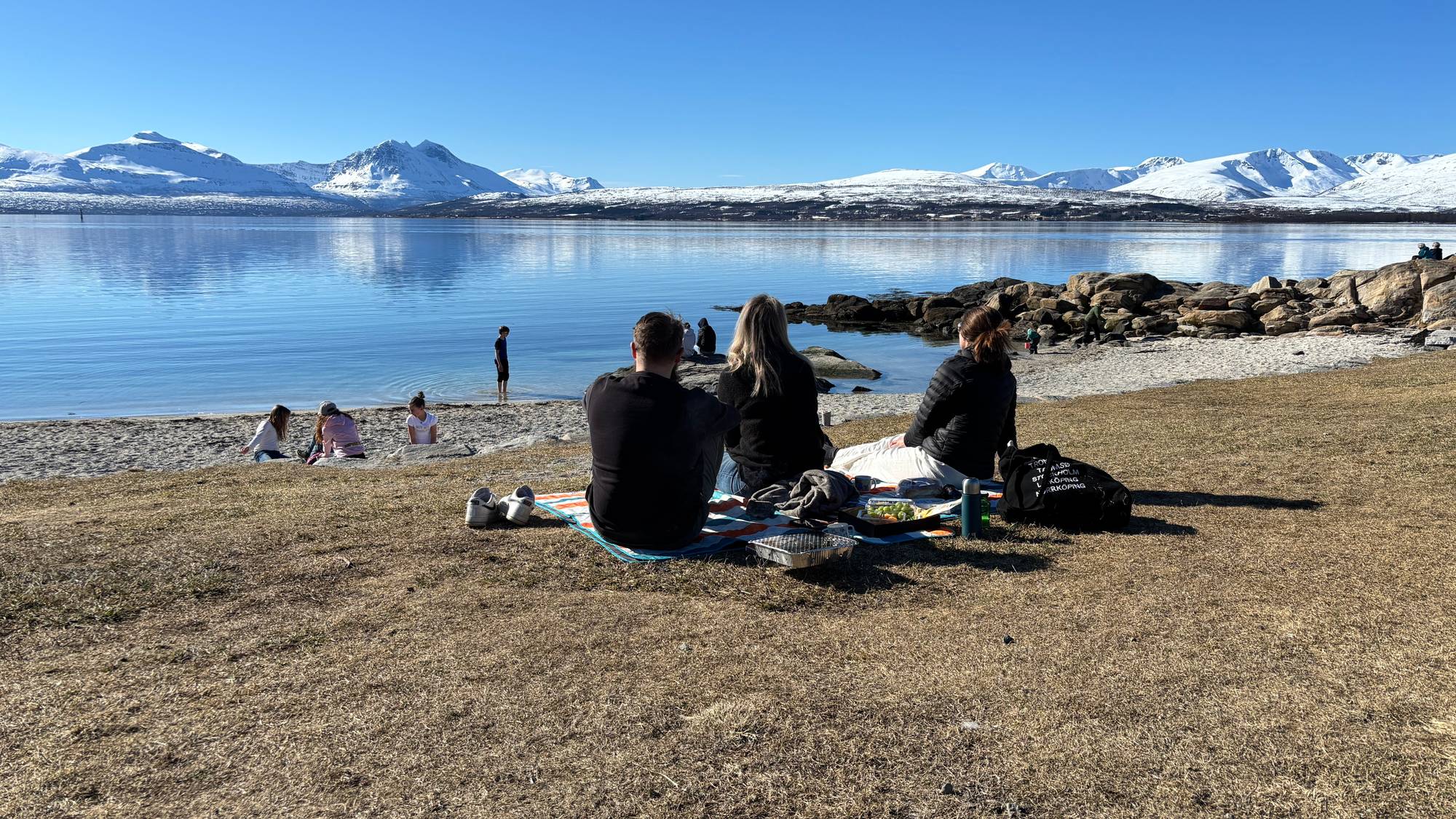 Mange mennesker samles for piknik ved fjorden. Flere barn leker på sandstranden i bakgrunnen. Snødekte fjell strekker seg i det fjerne under en klar blå himmel. Solen skinner og vannet ligger stille. (Bildebeskrivelsen er laget av en KI-tjeneste)