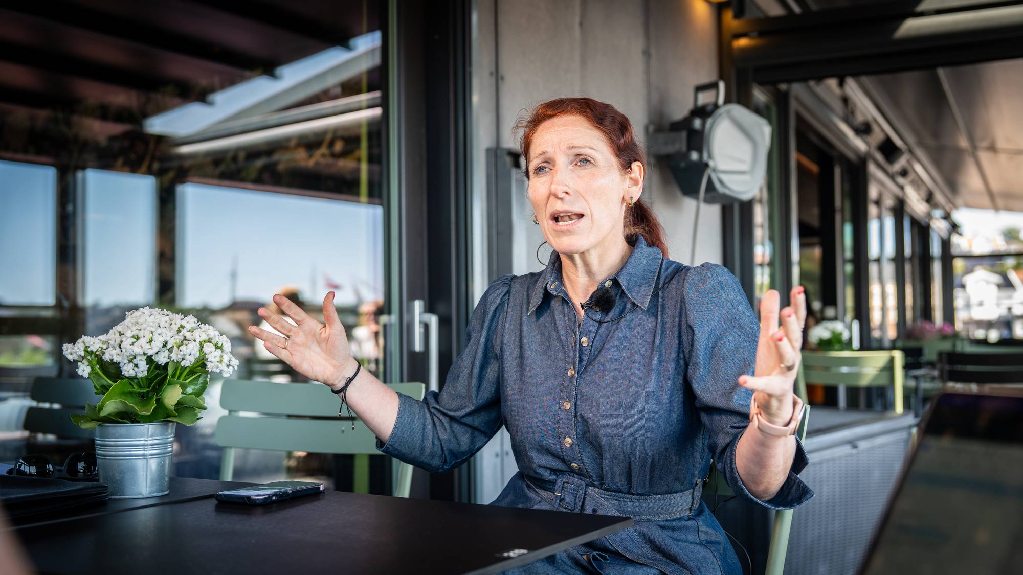 Kvinne i blå kjole gestikulerer mens hun snakker ved et bord utendørs. (Bildebeskrivelsen er laget av en KI-tjeneste)