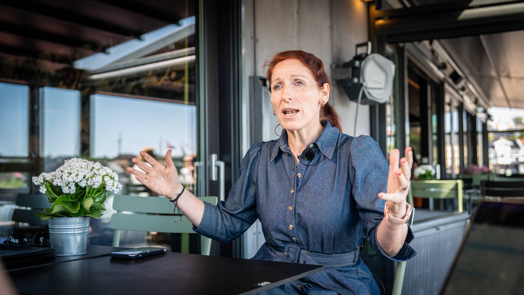 Kvinne i blå kjole gestikulerer mens hun snakker ved et bord utendørs. (Bildebeskrivelsen er laget av en KI-tjeneste)