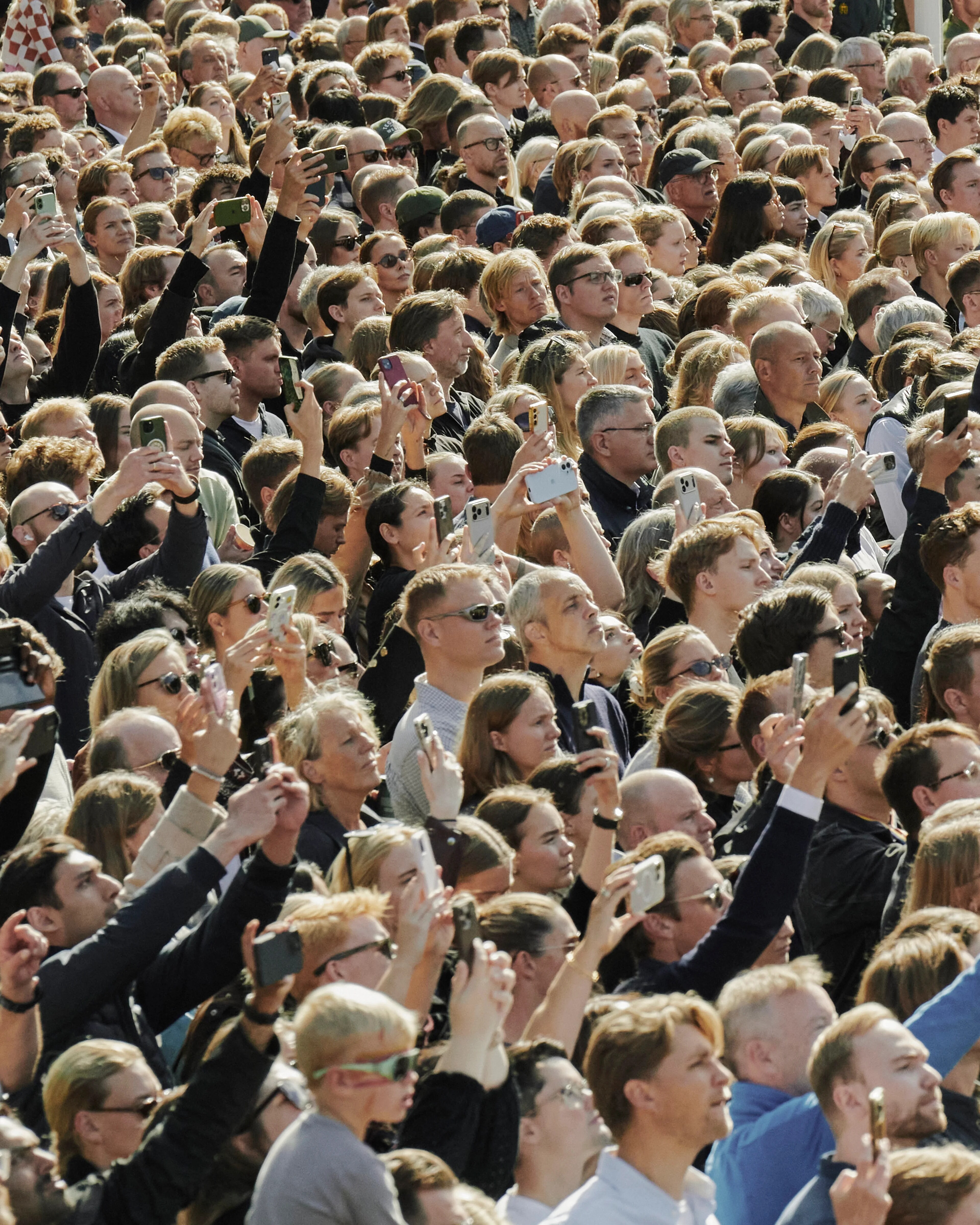 En stor folkemengde står utendørs og fotograferer med mobiltelefoner. I bakgrunnen står soldater. (Bildebeskrivelsen er laget av en KI-tjeneste)
