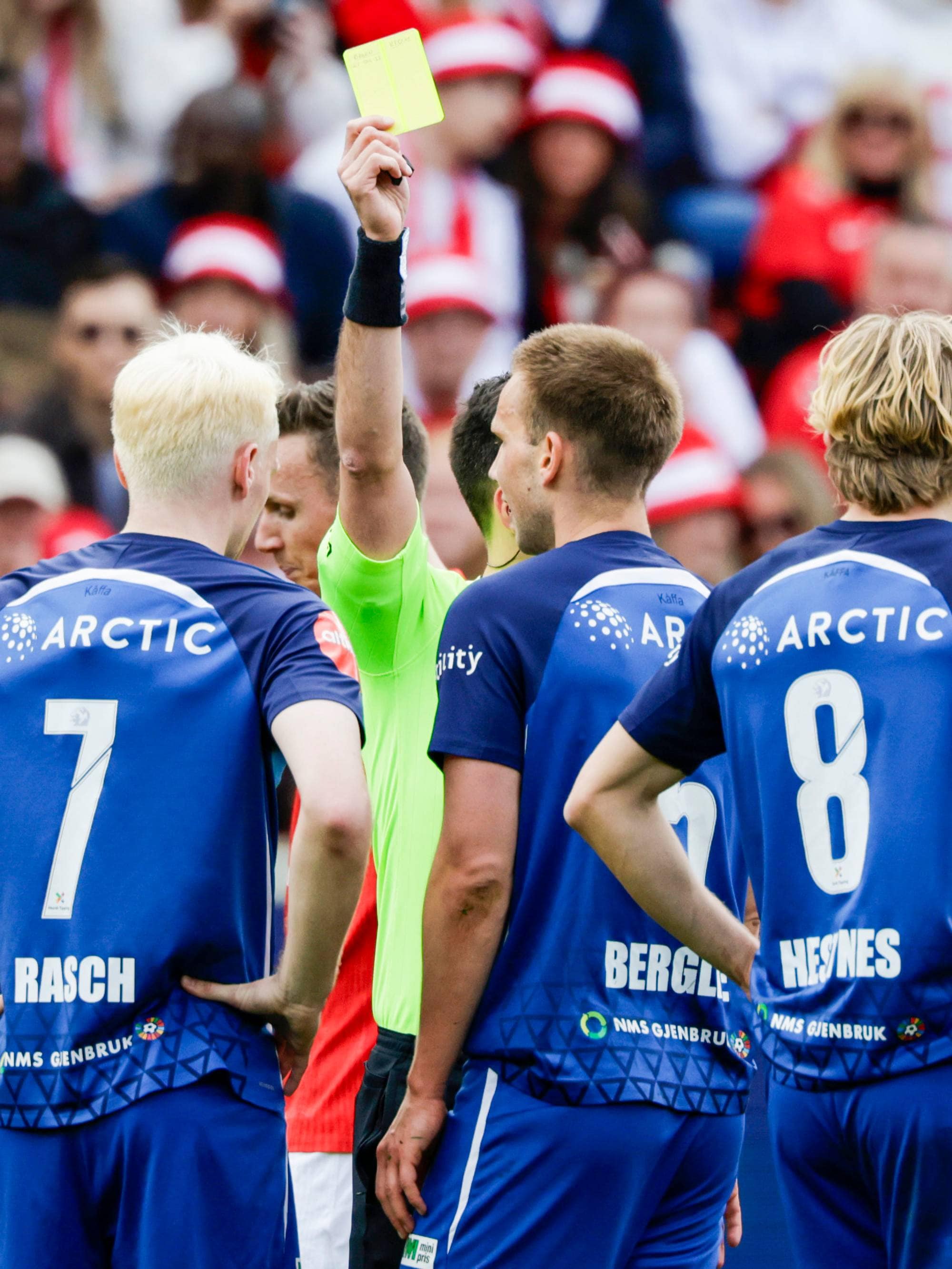 En fotballdommer gir et gult kort til en spiller. Andre spillere ser på. Tilskuere sitter på tribunen i bakgrunnen. (Bildebeskrivelsen er laget av en KI-tjeneste)