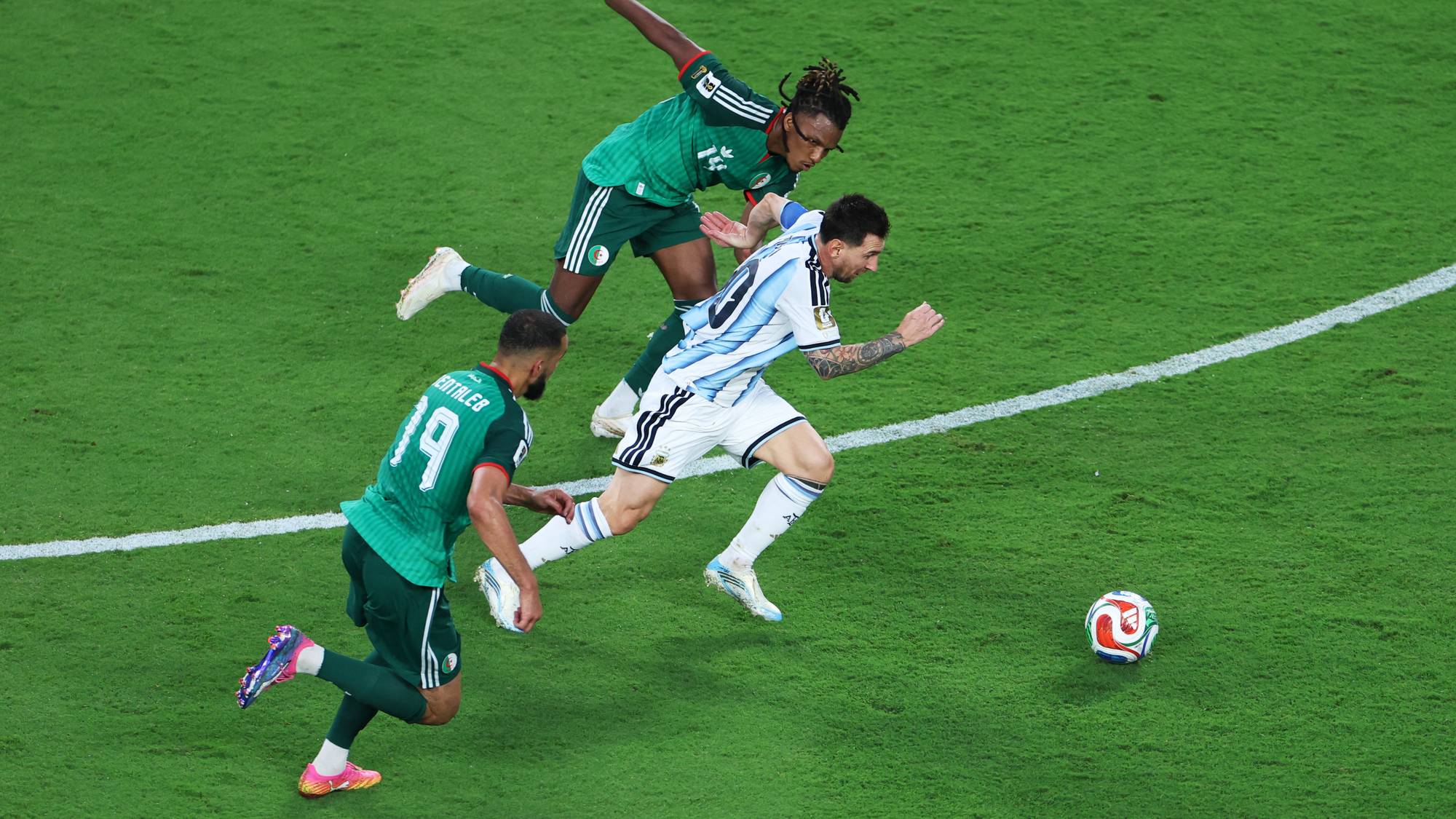 Tre fotballspillere er i luften under en kamp. En spiller i hvit drakt med blå striper, antatt å være Lionel Messi, hopper mens to spillere i grønn drakt prøver å hindre ham. En fotball ligger på gresset til høyre i bildet. Bildet er tatt fra et fugleperspektiv. (Bildebeskrivelsen er laget av en KI-tjeneste)