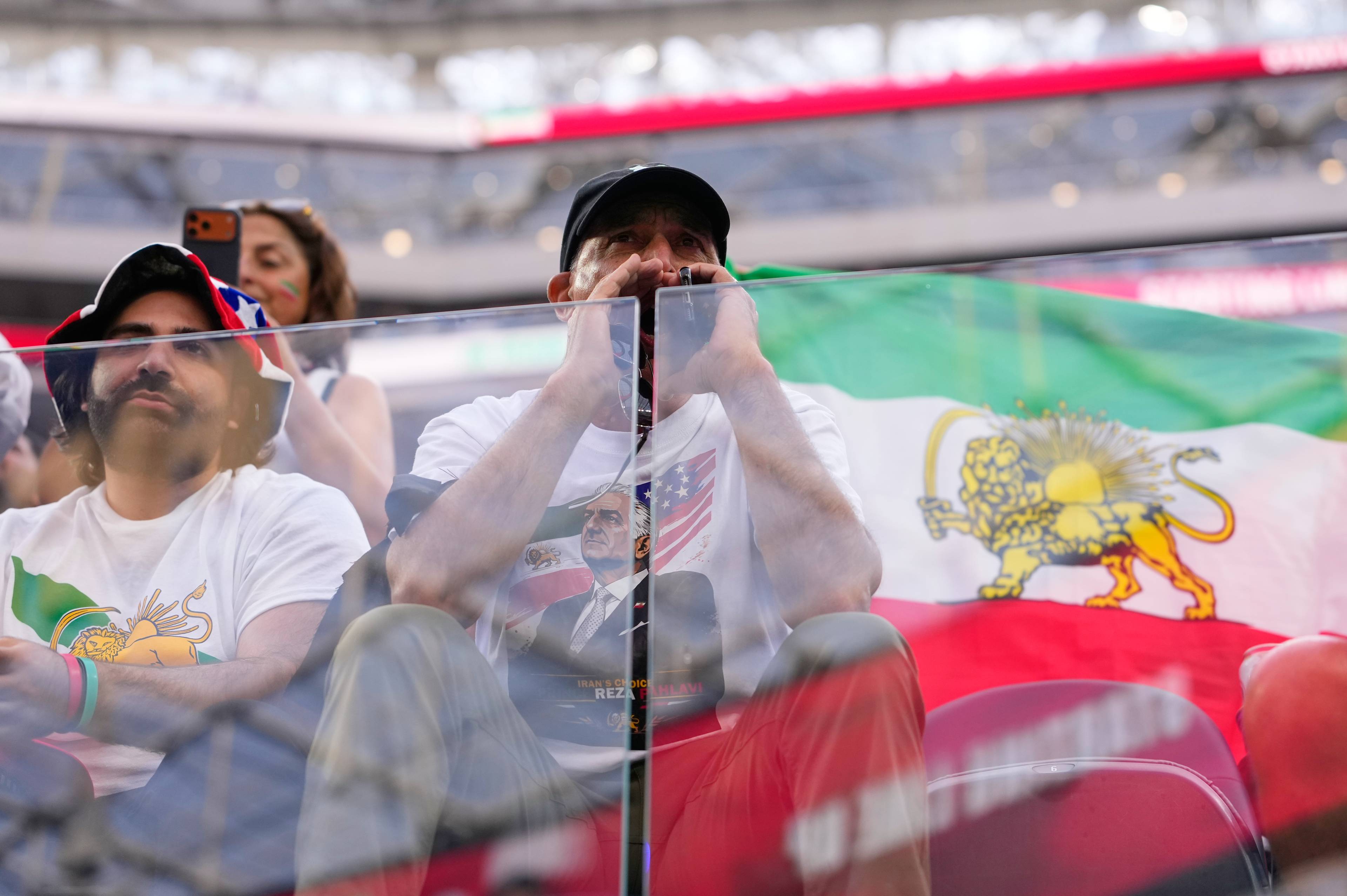 Menn sitter på tribunen med iranske flagg. En mann har en t-skjorte med bilde av Reza Pahlavi og amerikansk flagg. En annen mann har på seg en hatt med stjerner og striper. Bakgrunnen viser tribuner og iranske flagg. (Bildebeskrivelsen er laget av en KI-tjeneste)