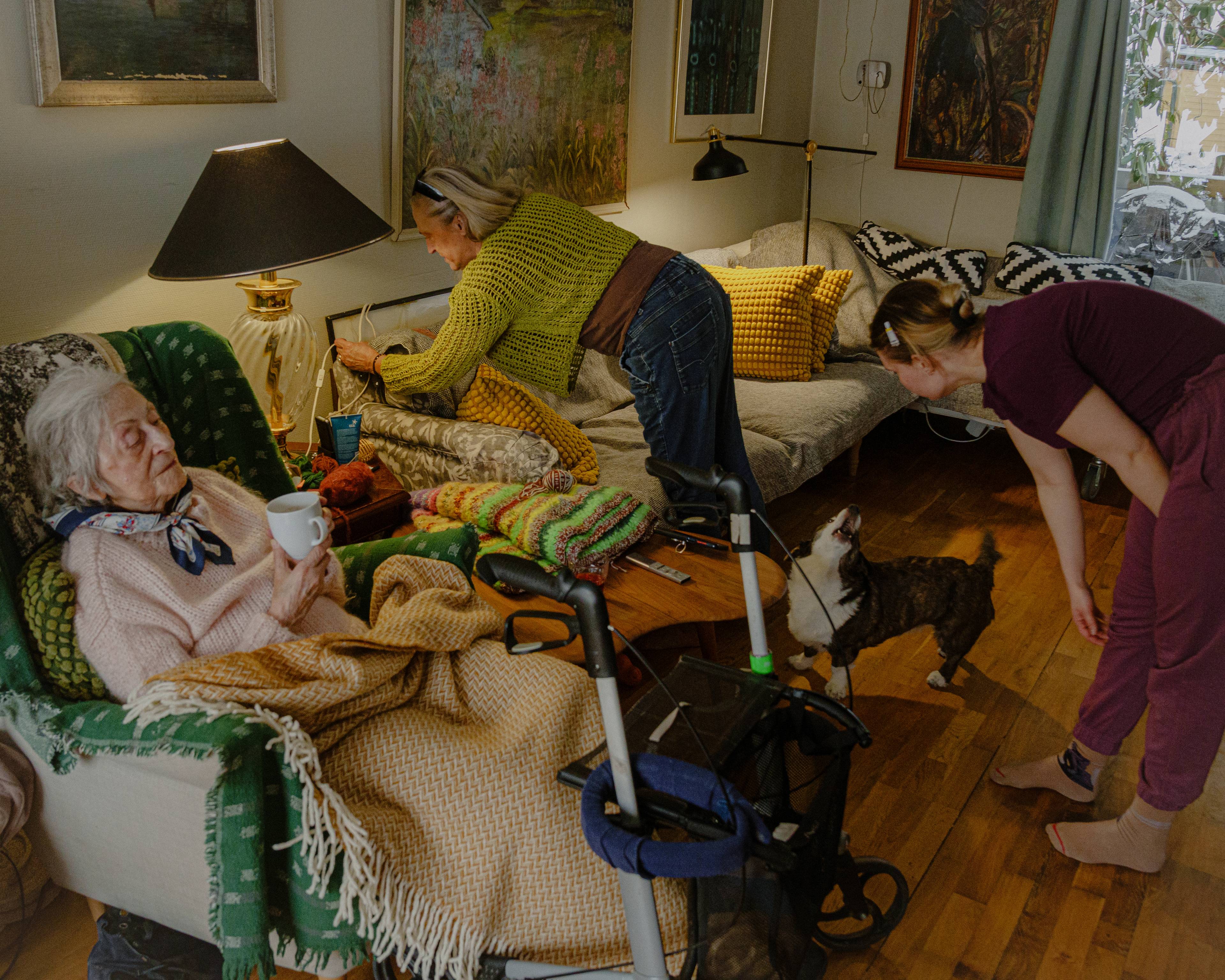 Magdalena sitter i lenestolen sin med en kopp kaffe. Ved siden av skrur Edith på en lampe, mens Celina ser ned mot en hund