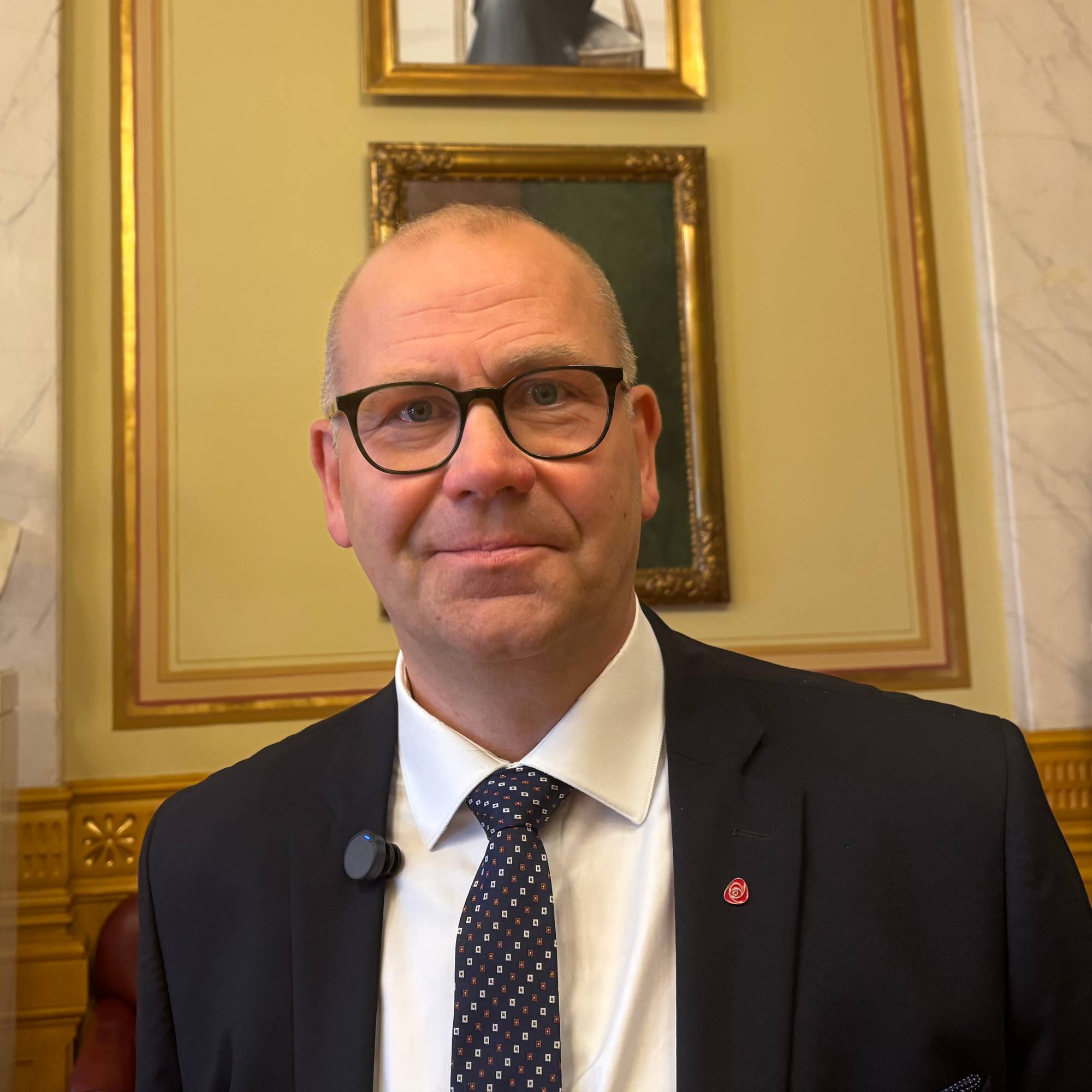 En mann i dress står i et rom med detaljerte vegger og bølgende tak. I bakgrunnen er det malerier og skulpturer. Mannen har mikrofonclip og ser mot kameraet. Rommet har treverk og marmor som gir det et formelt preg. (Bildebeskrivelsen er laget av en KI-tjeneste)