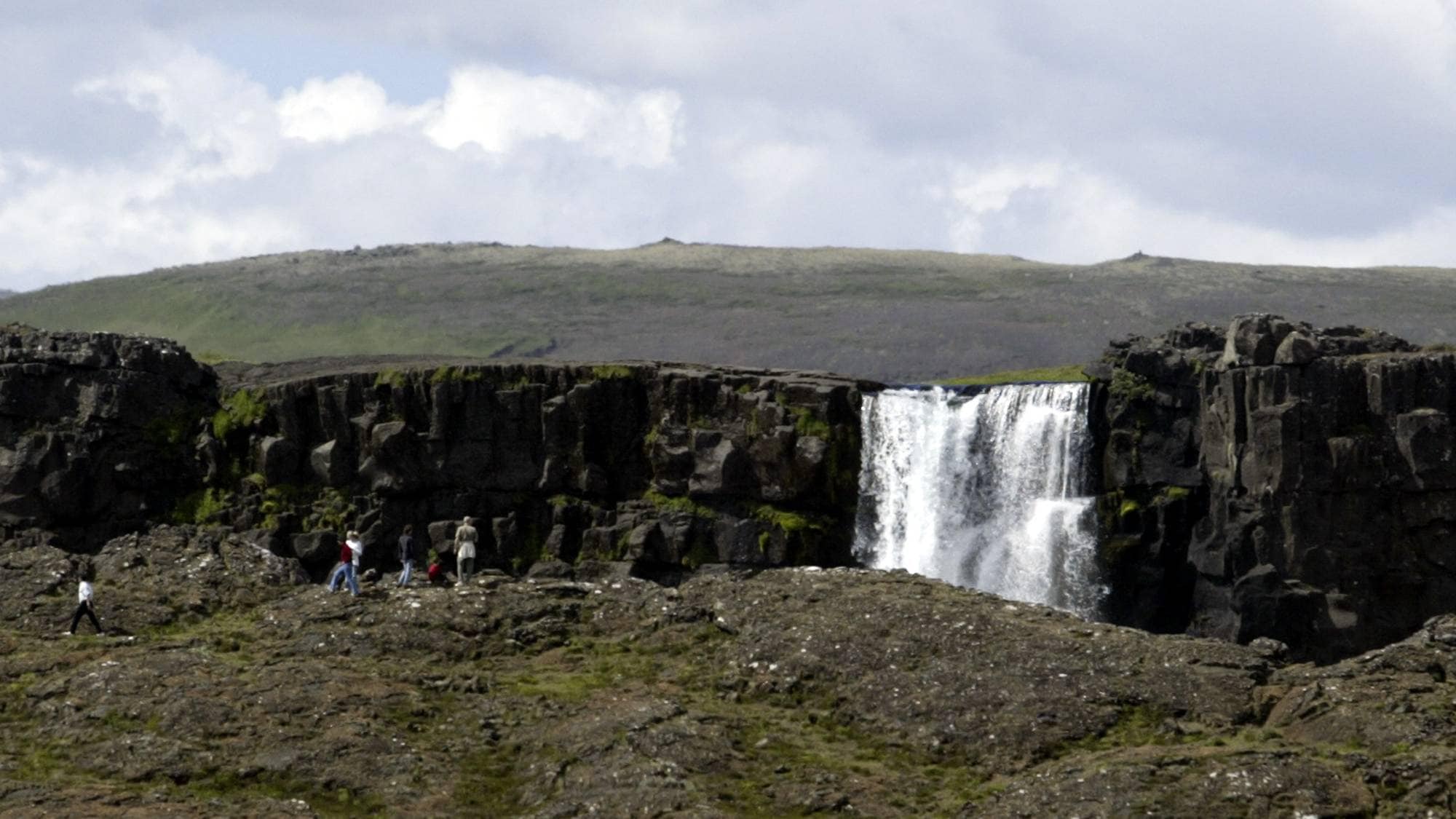 Turister går ved en foss i et steinete landskap i Þingvellir nasjonalpark på Island. (Bildebeskrivelsen er laget av en KI-tjeneste)