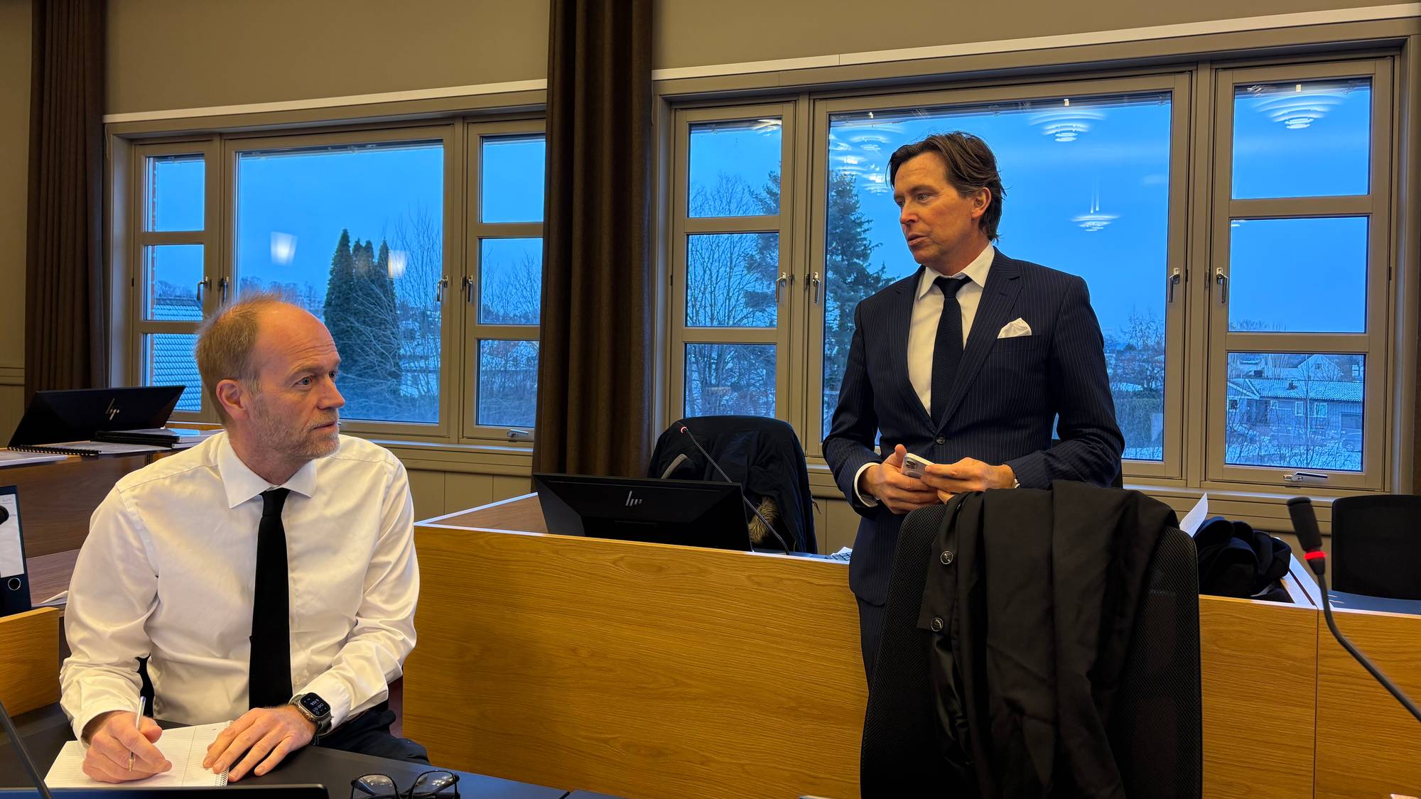 En mann i hvit skjorte og slips sitter på en stol i en rettssal i Agder lagmannsrett. Ved siden av står en mann i mørk dress.