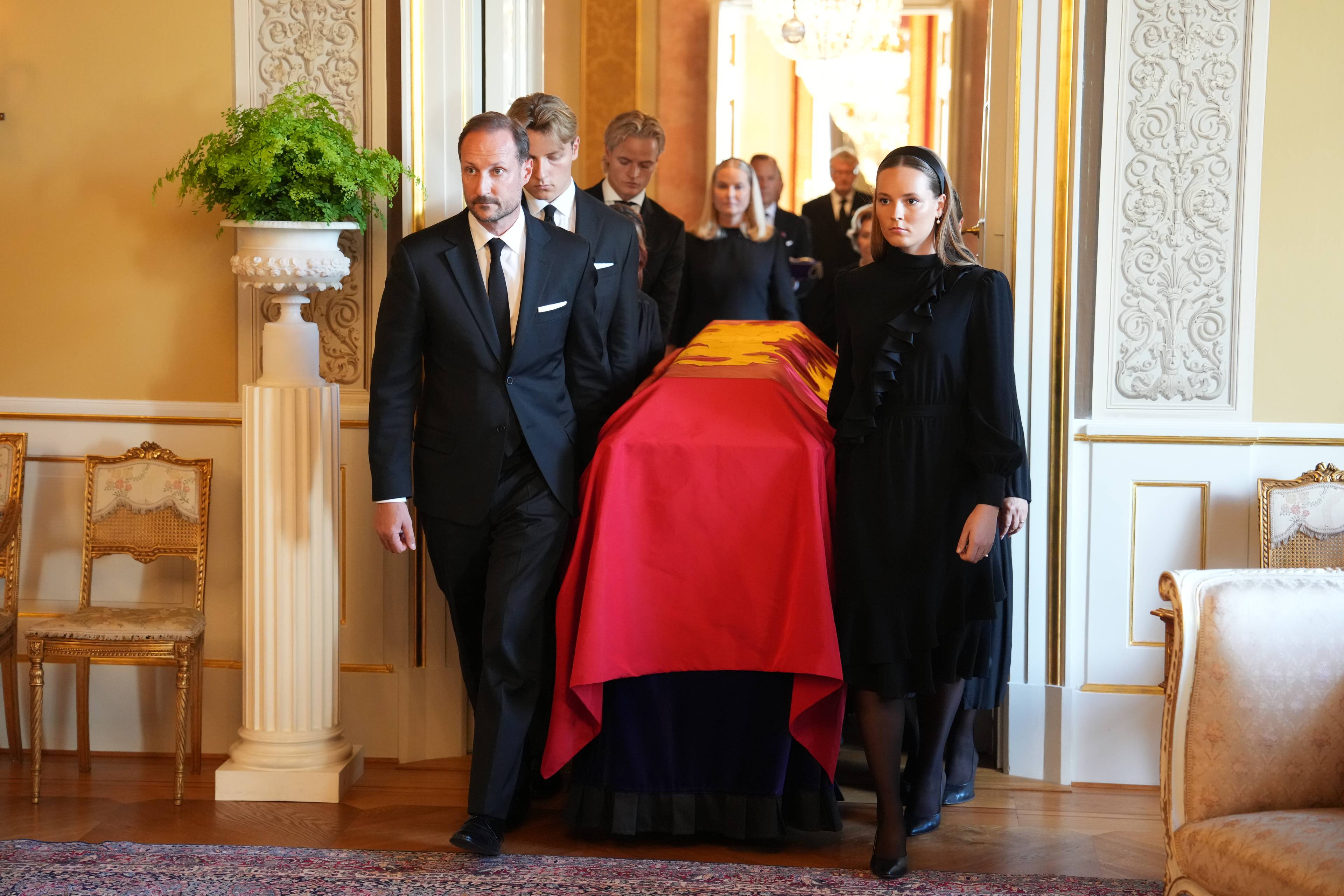 Kronprins Haakon og prinsesse Ingrid Alexandra bærer en kiste dekket av et rødt flagg i et slott. (Bildebeskrivelsen er laget av en KI-tjeneste)