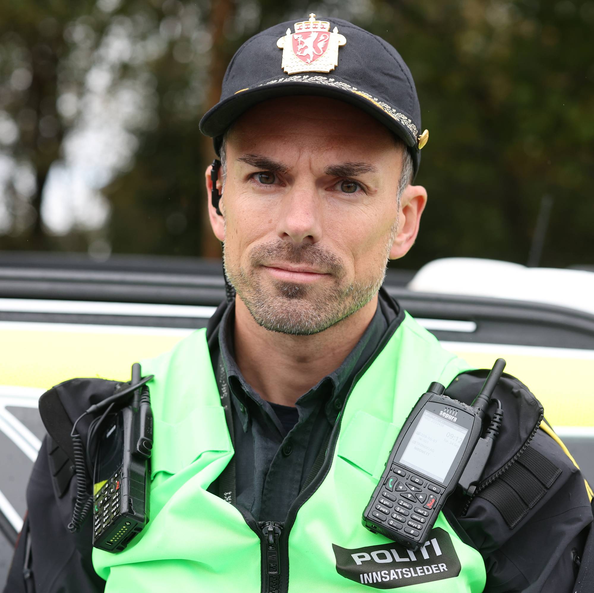 Innsatsleder fra politiet med gul vest foran en politibil. (Bildebeskrivelsen er laget av en KI-tjeneste)