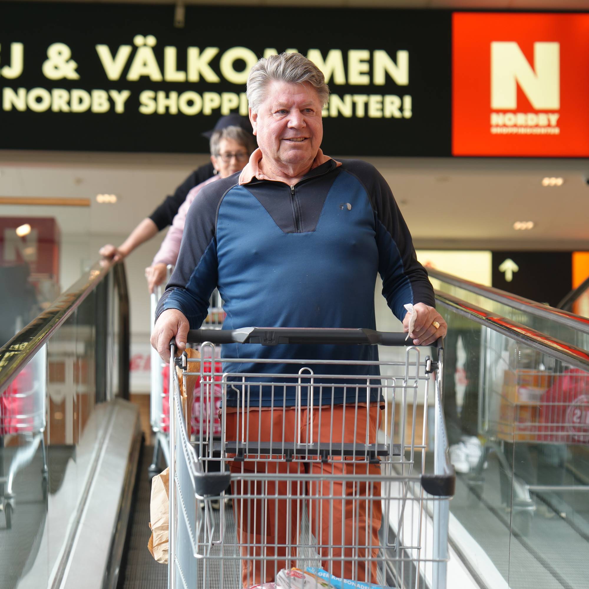 En mann med handlevogn på rulletrapp. Han har på seg en blå genser og oransje bukser. Bak ham er en kvinne med en handlevogn. I bakgrunnen er det et skilt der det står «HEJ & VÄLKOMMEN TILL NORDBY SHOPPING CENTER». (Bildebeskrivelsen er laget av en KI-tjeneste)