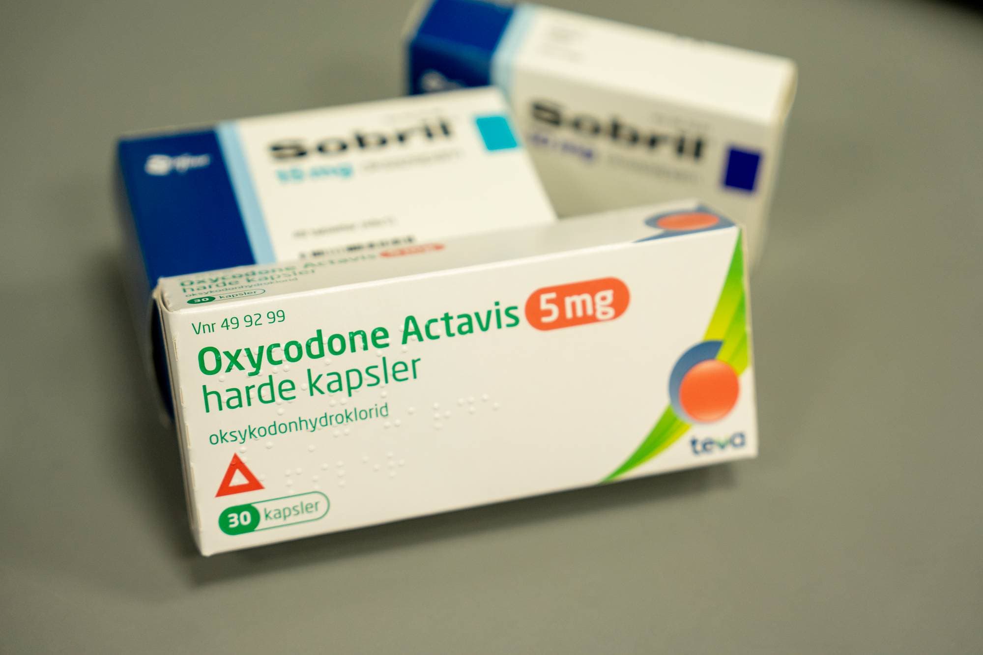 Bokser med medisinene Oxycodone Actavis og Sobril er plassert på et grått underlag. Oxycodone Actavis 5mg er i forgrunnen med teksten "harde kapsler" og "oksykodonhydroklorid" synlig. Bak ligger to esker med Sobril. (Bildebeskrivelsen er laget av en KI-tjeneste)