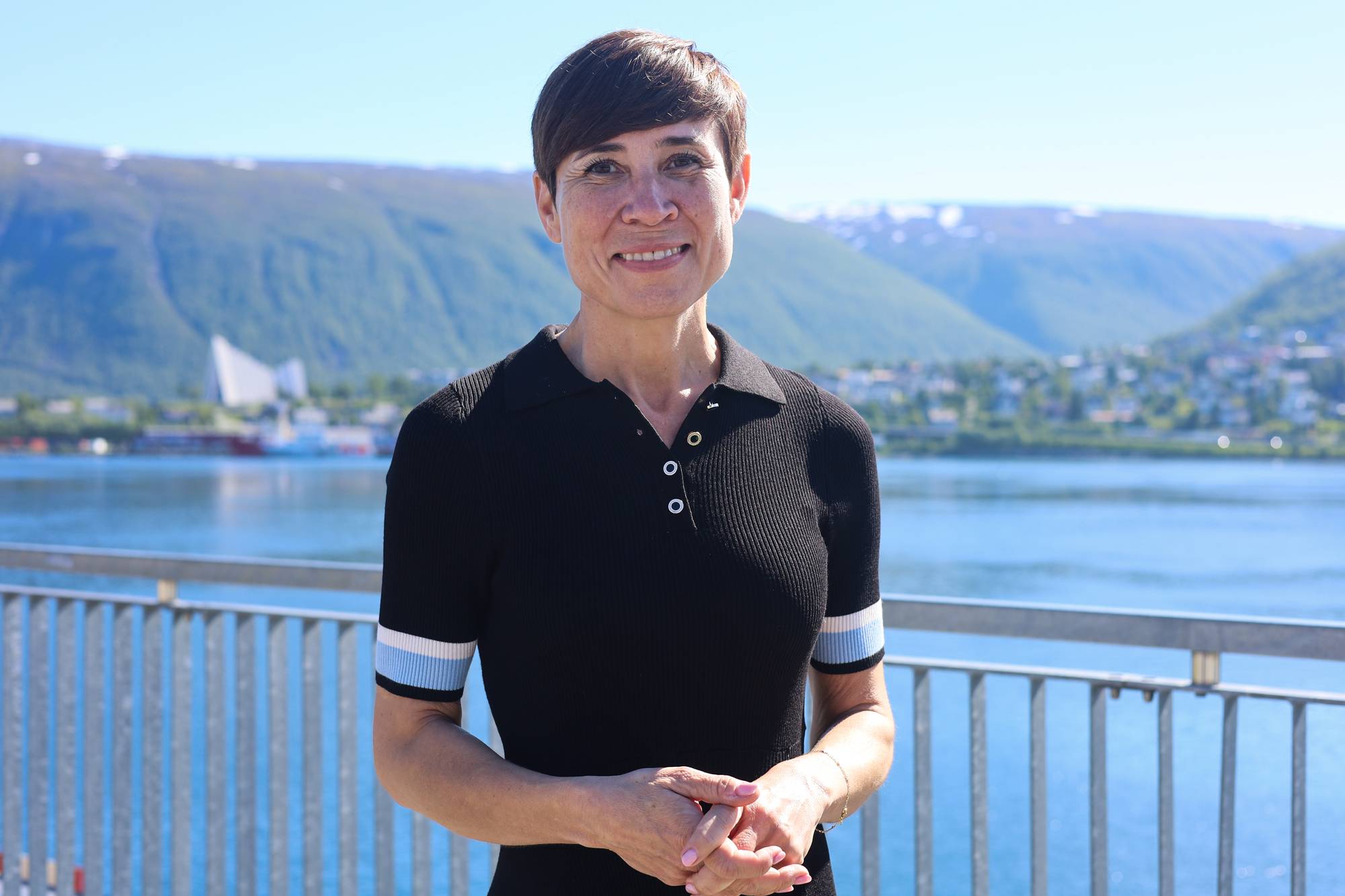 En kvinne med kort sort hår står utendørs. Ishavskatedralen i Tromsø vises i bakgrunnen.