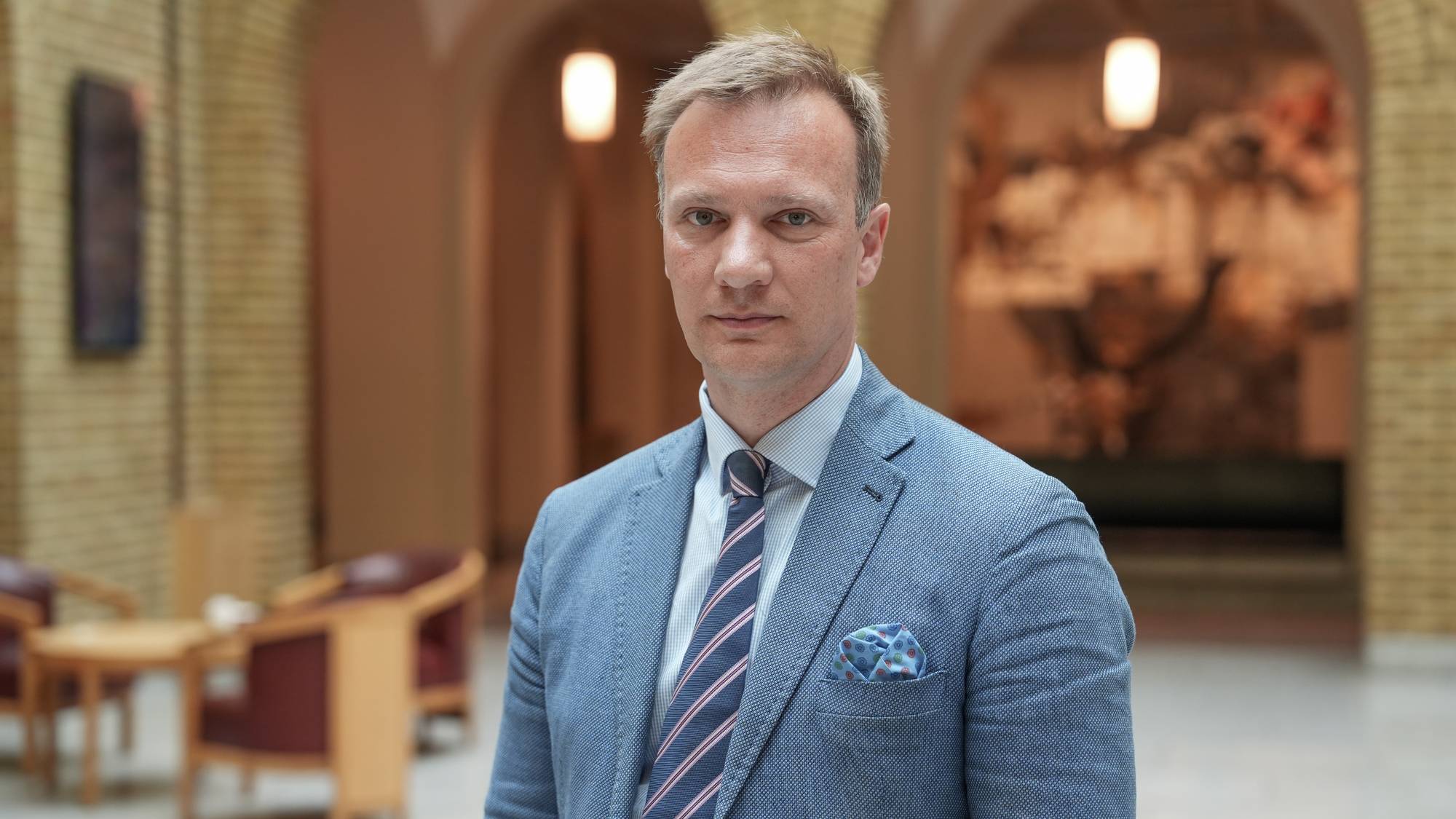 Mann i dress og stripete slips står foran murvegger i et innendørs lokale. (Bildebeskrivelsen er laget av en KI-tjeneste)