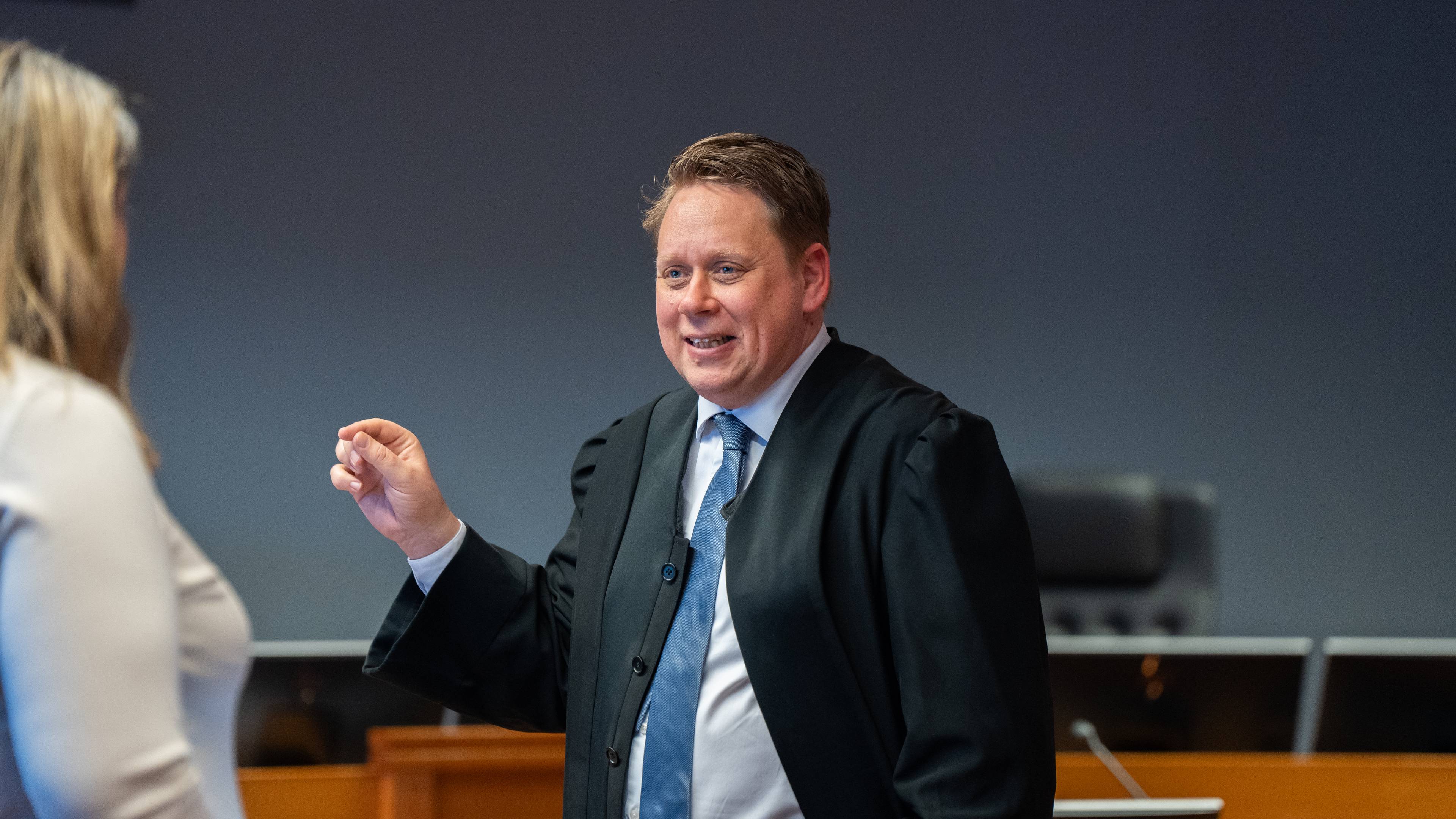 En mann i dommerdrakt snakker til en kvinne. Mannen gestikulerer med venstre hånd og peker med høyre hånd. Bak dem er det en mørk vegg og møbler.