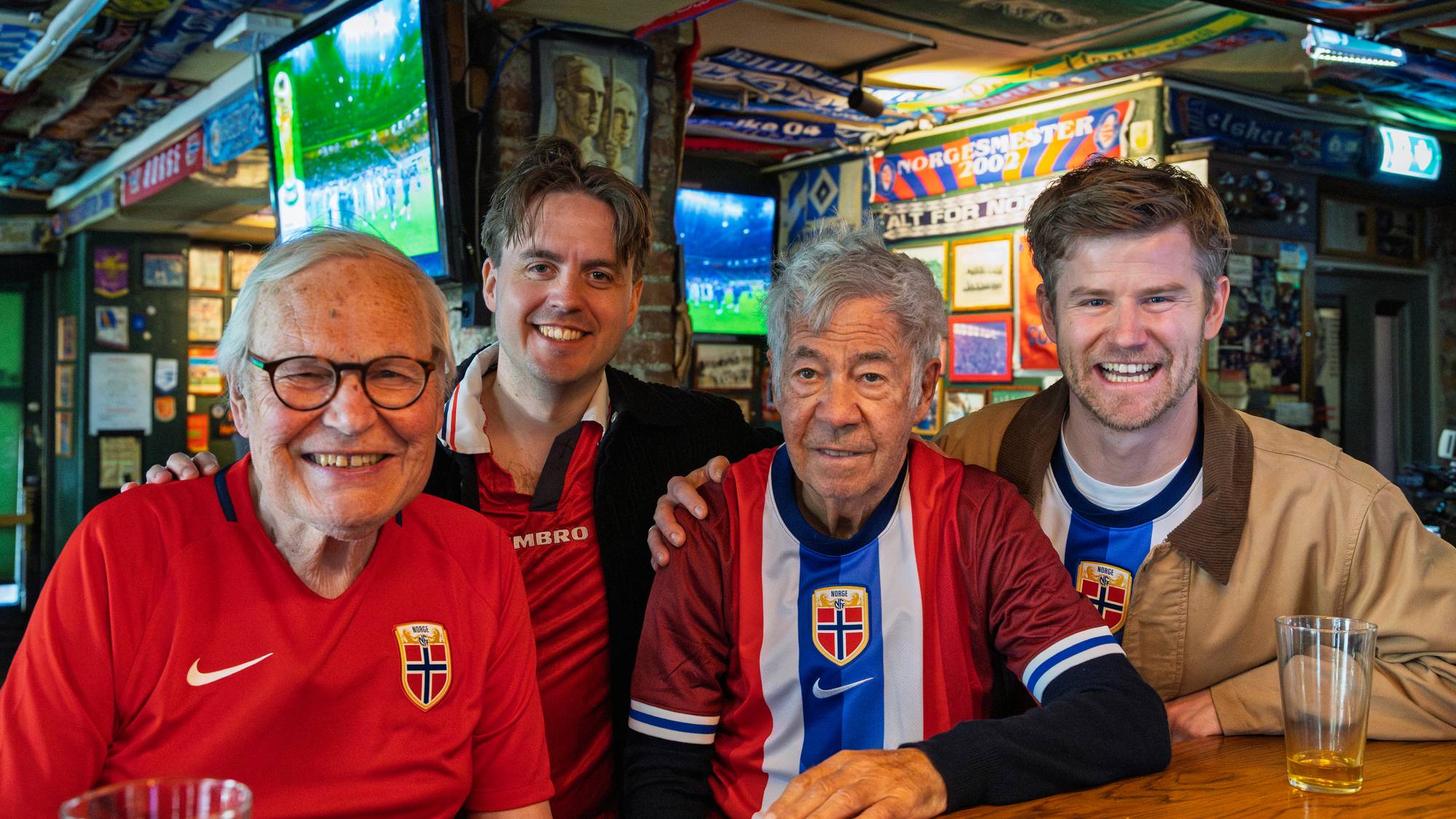 Fire menn smiler mot kameraet i en bar. Tre av dem har på seg fotballdrakter med norske flagg. Bak dem henger flere fotballdrakter og skjerf, og det er en TV på veggen som viser en fotballkamp. 