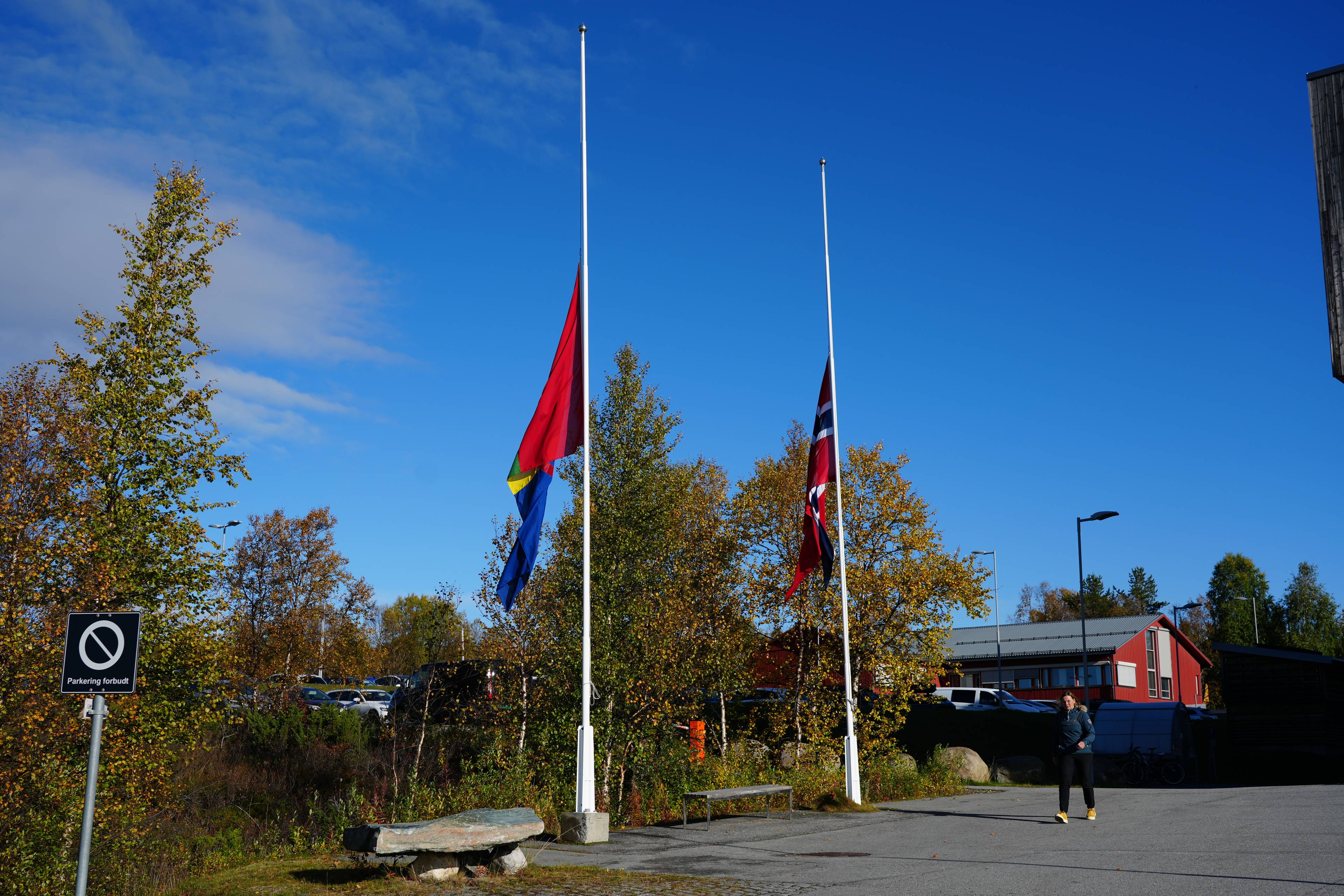 Norsk flagg og et annet flagg på halv stang utenfor en bygning. Kvinne går på en asfaltert plass. (Bildebeskrivelsen er laget av en KI-tjeneste)