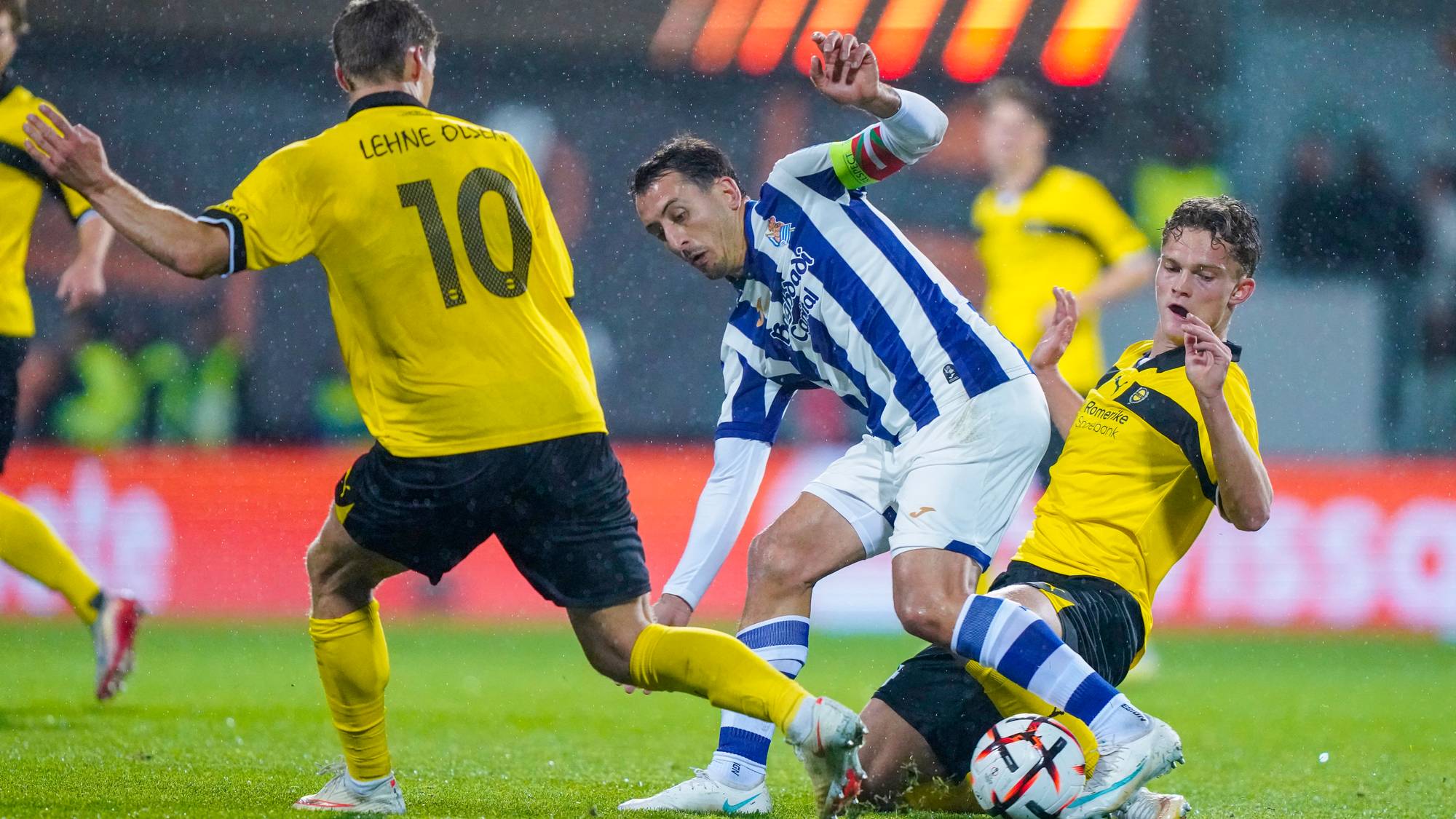 Fotballspillere i duell om ballen på en gressbane under en kamp. (Bildebeskrivelsen er laget av en KI-tjeneste)