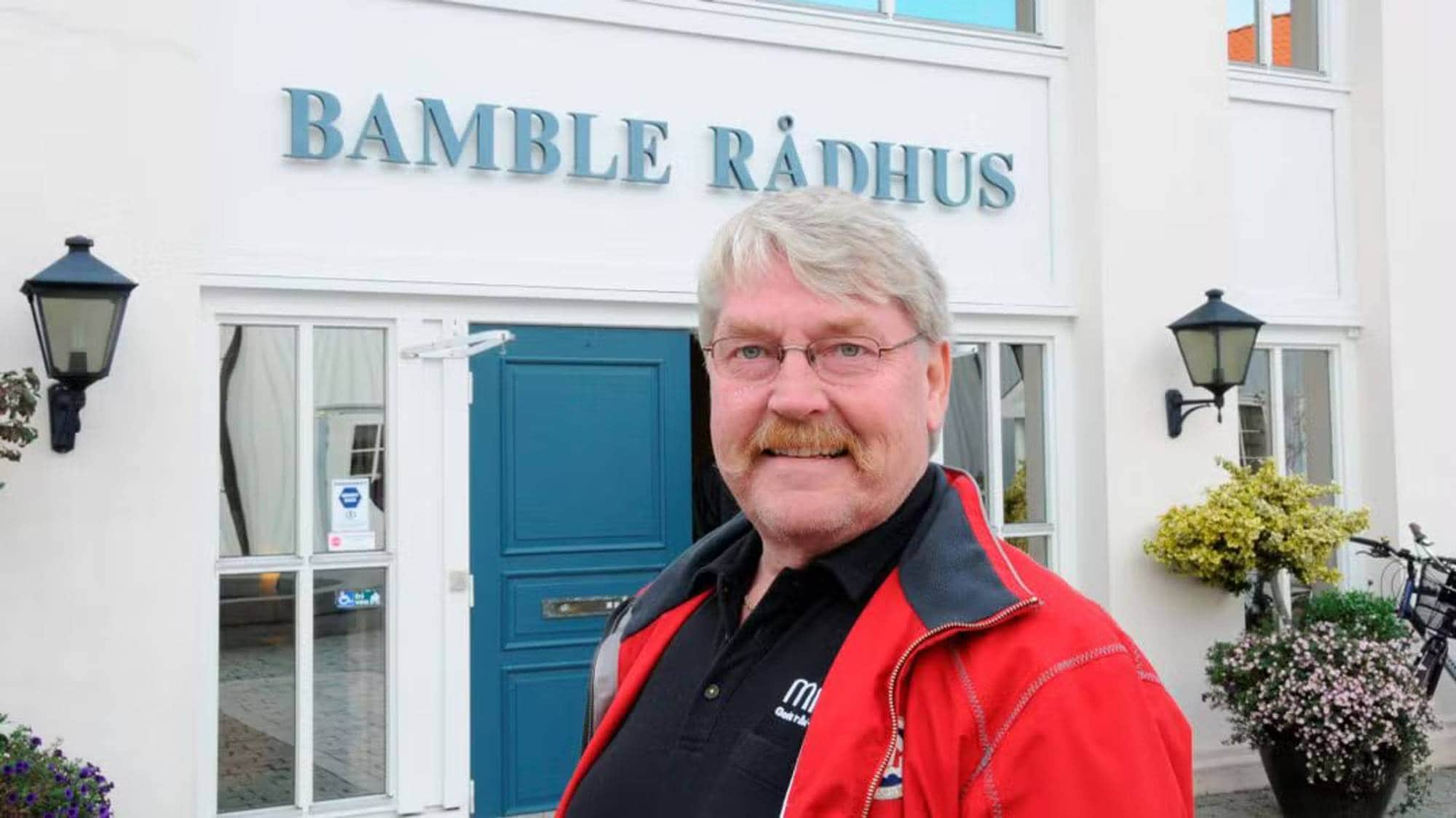 En mann med rød jakke smiler til kamera, foran rådhuset i Bamble. 
