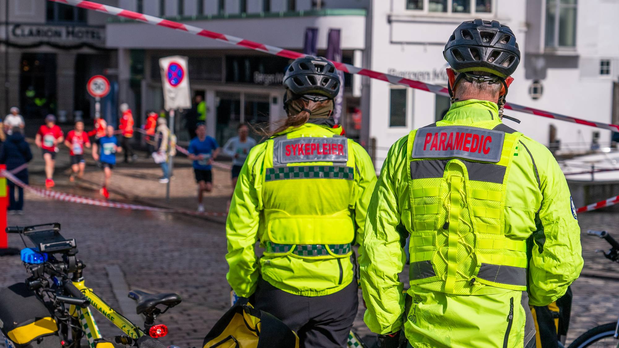 To helsepersonell i gule jakker med ryggen til, i eit gatekryss langs løypa på Bergen City Marathon 2026.