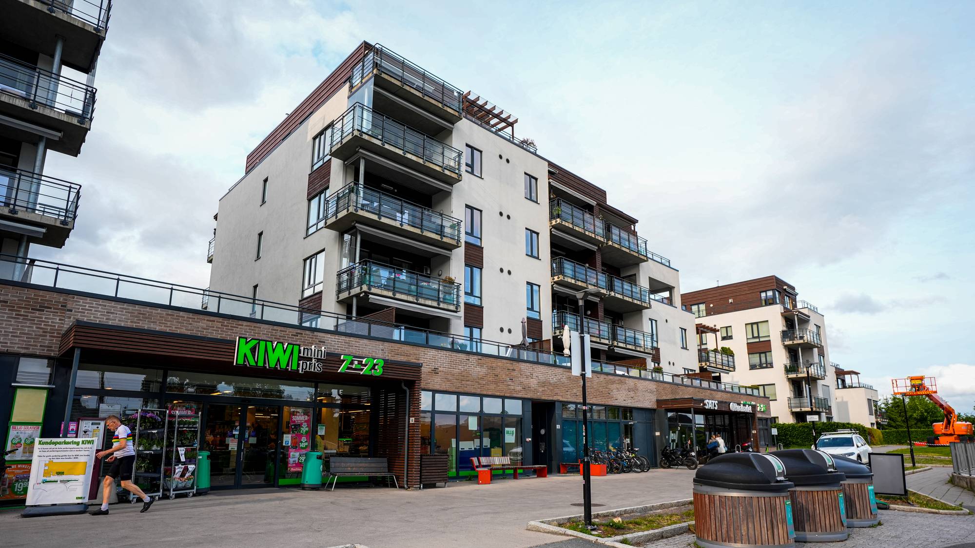 Boligblokk med en Kiwi-butikk i første etasje og en person som går på fortauet utenfor. (Bildebeskrivelsen er laget av en KI-tjeneste)