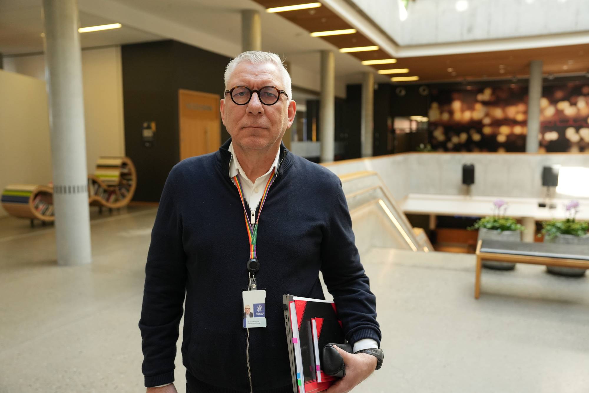 En mann med grått hår og mørke briller står i en stor hall. Han har et nøkkelkort rundt halsen og permer og pc under armen. 