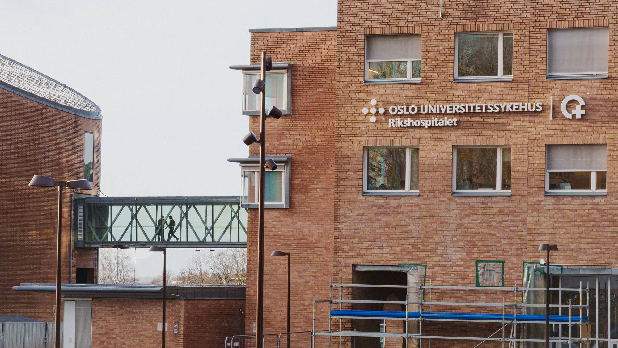 Oslo Universitetssykehus Rikshospitalet er vist med en glassbro. To personer går på broen mellom to bygninger. En byggeplass med stillas er synlig. Flere elsparkesykler er parkert. (Bildebeskrivelsen er laget av en KI-tjeneste)