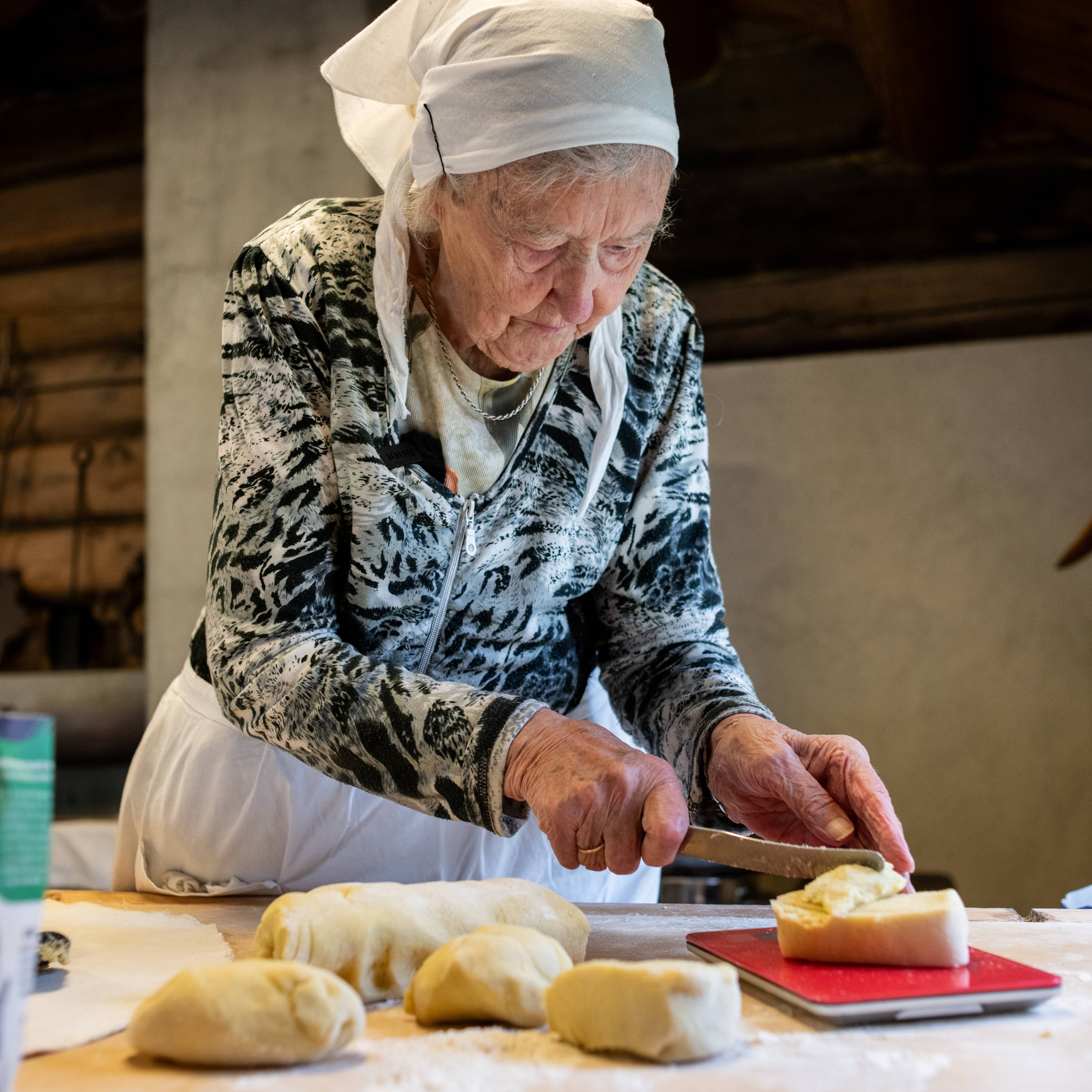 Eldre kvinne med hodeplagg skjærer deigstykker på et kjøkken.