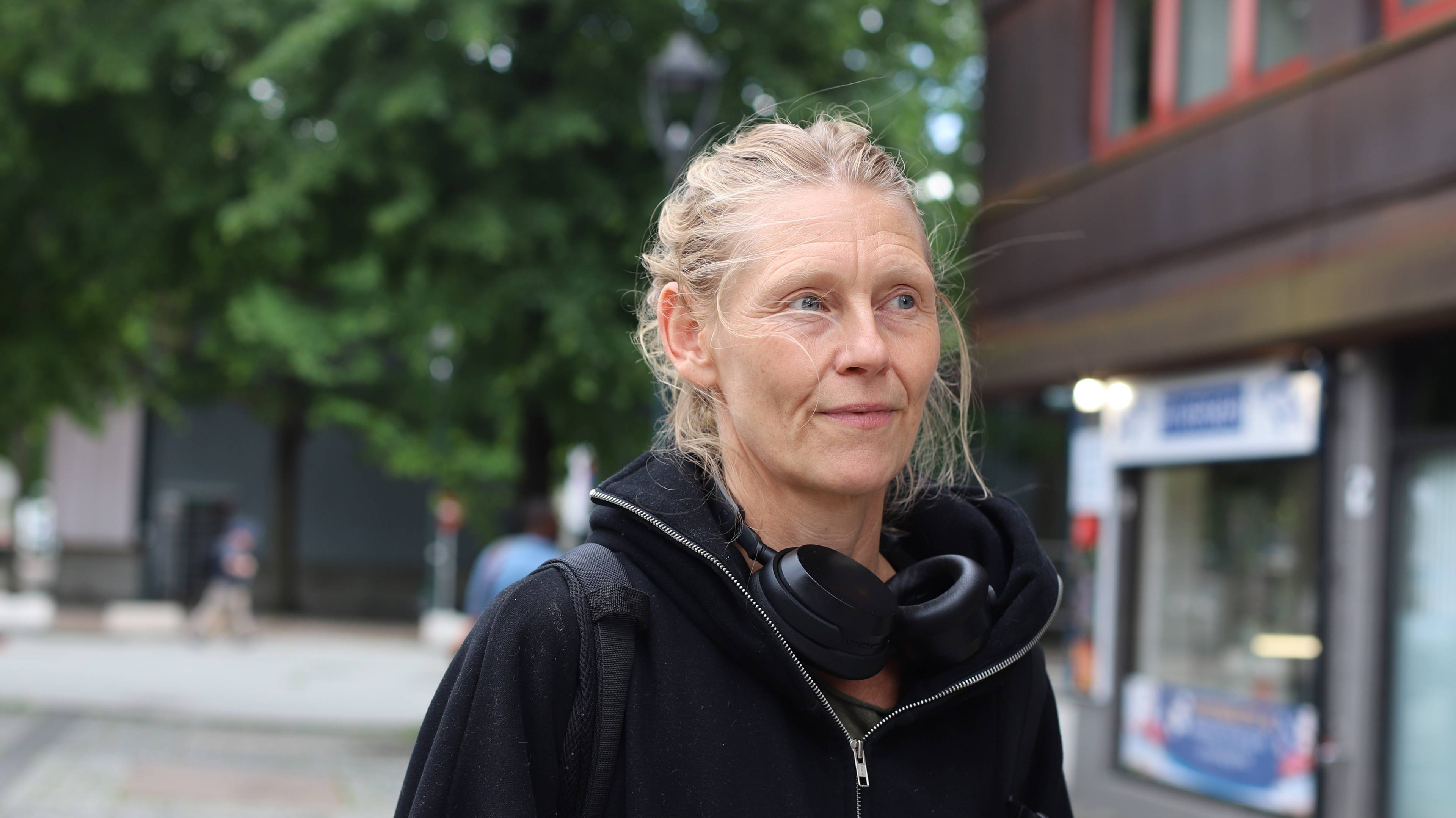 En kvinne med sort hettegenser og headset rundt halsen og blondt hår står ute på en gate og ser bort,