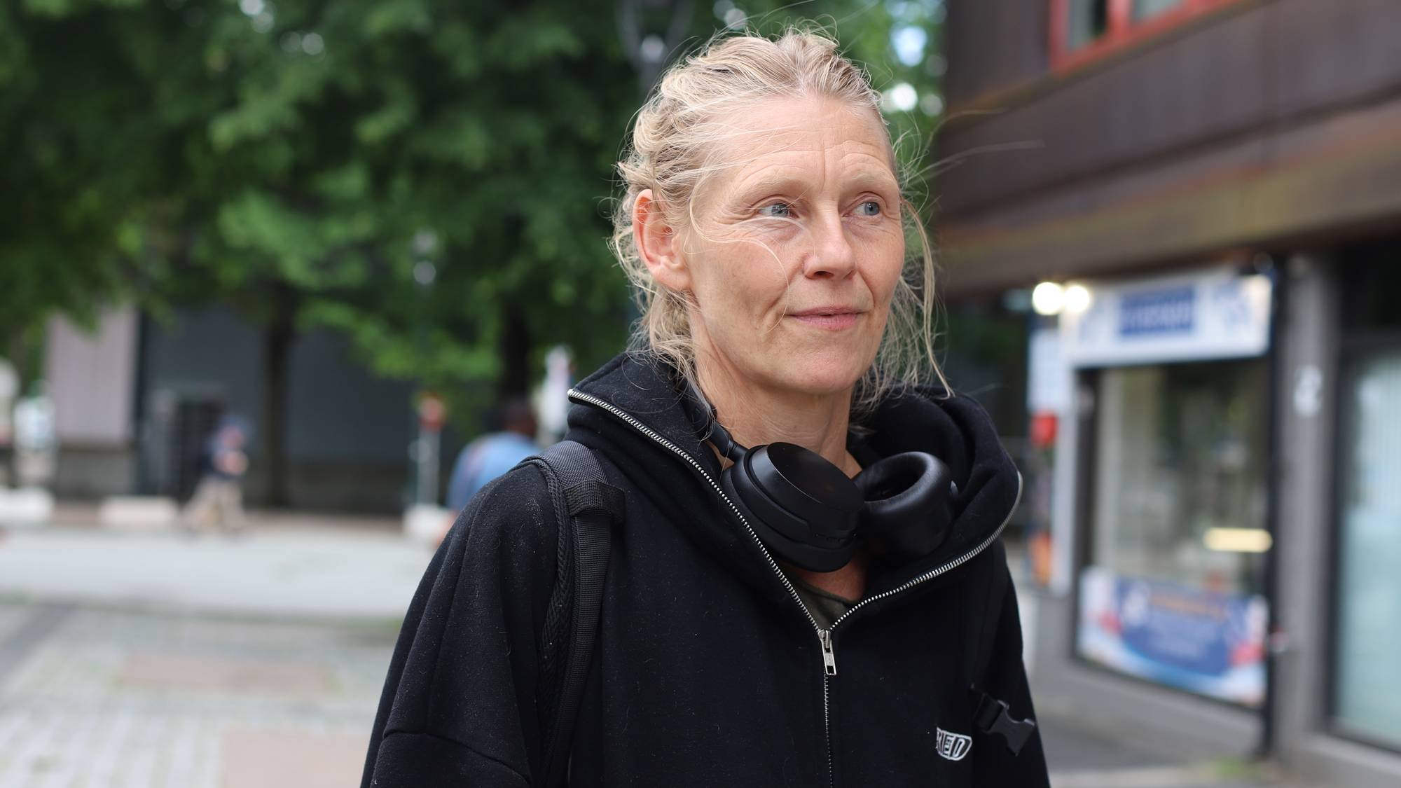 En kvinne med sort hettegenser og headset rundt halsen og blondt hår står ute på en gate og ser bort,