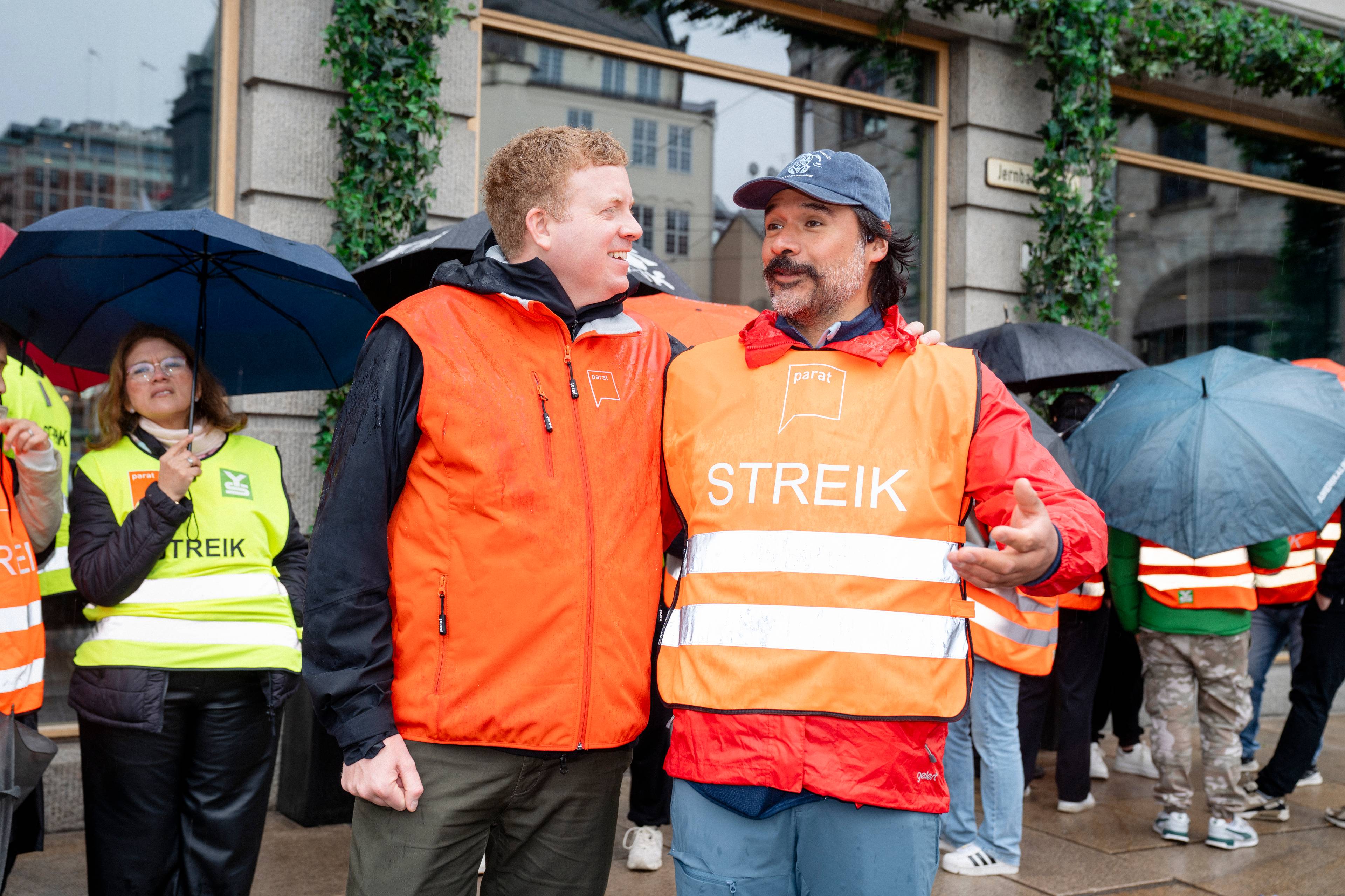 To menn i oransje vest med påskriften "STREIK" står sammen. De er en del av en gruppe mennesker som står ute i regnvær, mange med paraplyer. Bakgrunnen viser en bygate med bygninger. (Bildebeskrivelsen er laget av en KI-tjeneste)