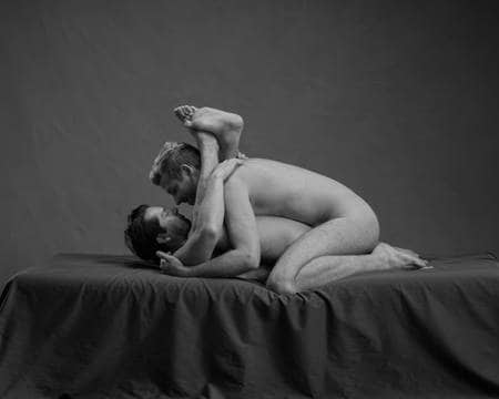 En naken mann med mørkt hår ligger på ryggen med bena bretta opp rundt halsen på en annen naken mann som ligger over han