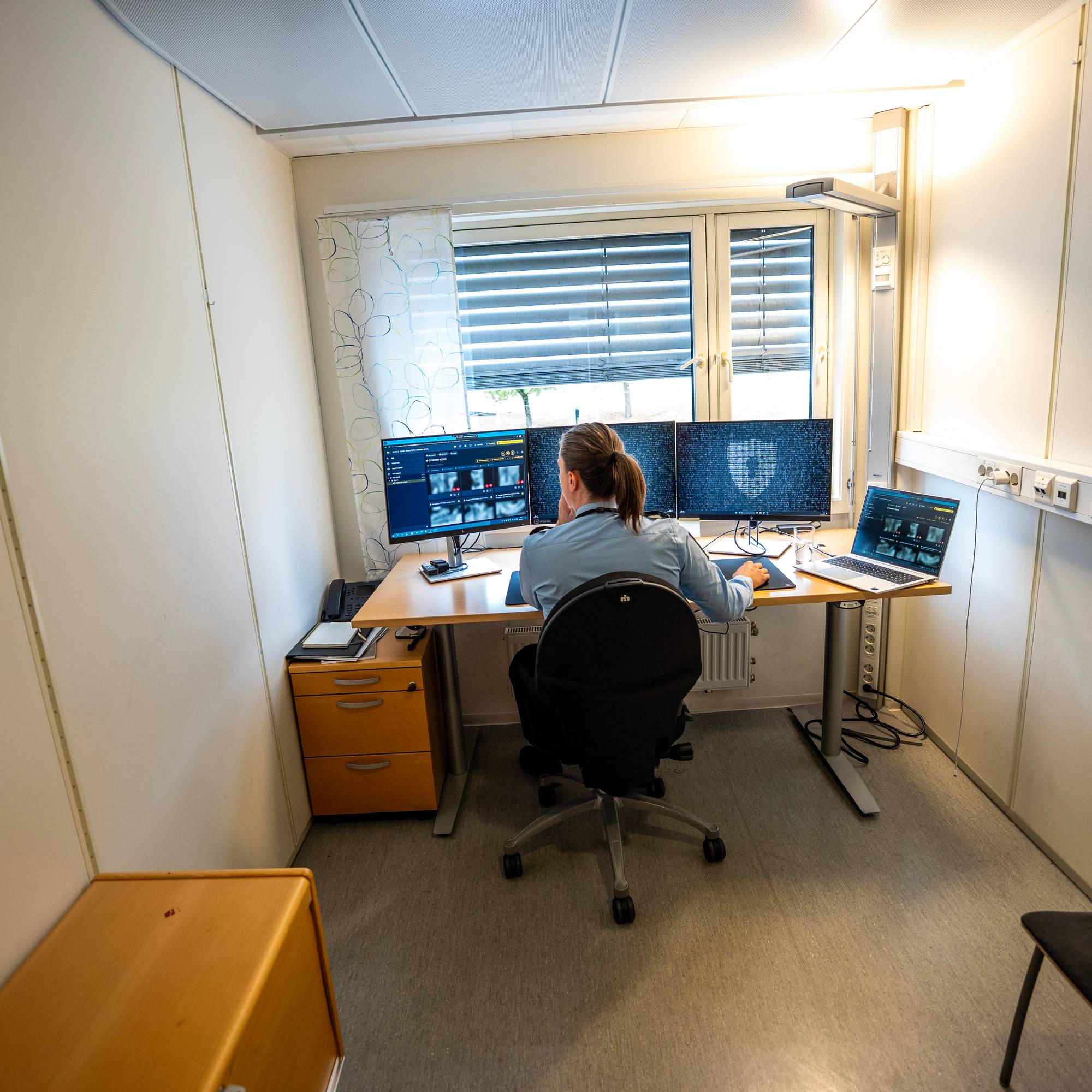 En politiansatt sitter ved et skrivebord på et kontor og arbeider foran flere dataskjermer. (Bildebeskrivelsen er laget av en KI-tjeneste)