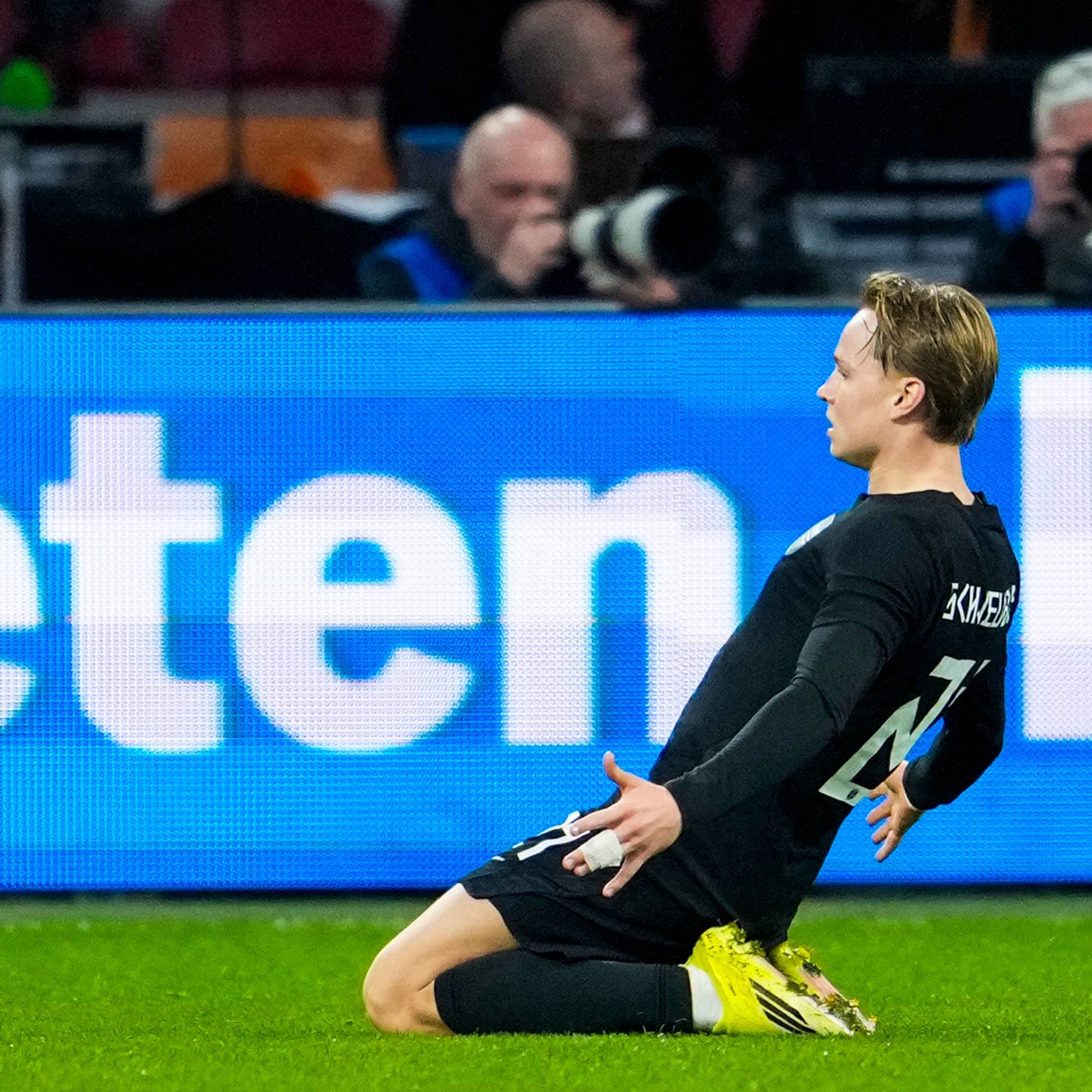En fotballspiller i svart drakt feirer et mål på en grønn gressbane. Spilleren er på kne og ser til siden. Bak ham vises en blå reklame med store hvite bokstaver. (Bildebeskrivelsen er laget av en KI-tjeneste)