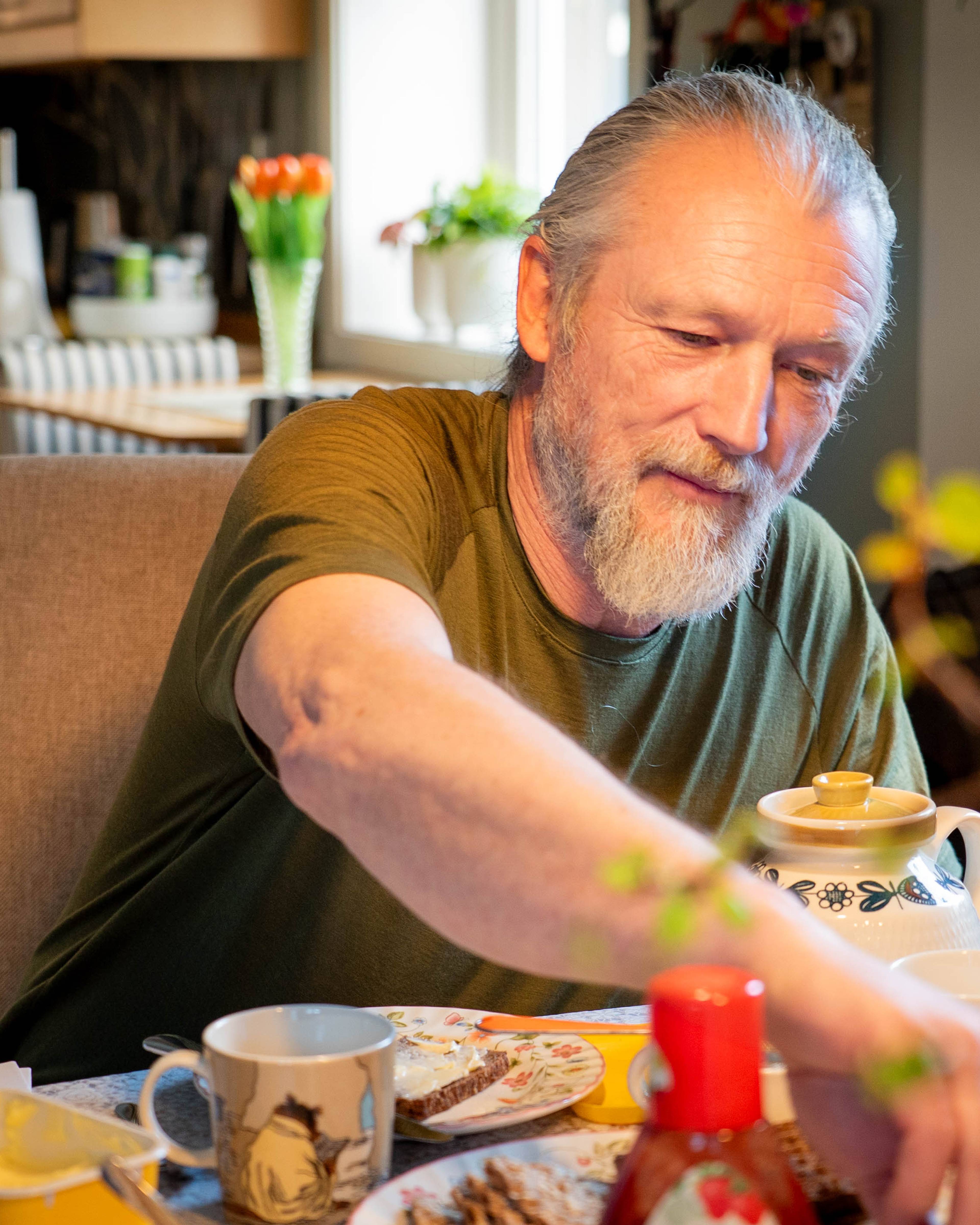 En mann med grått hår i hestehale som sitter ved et spisebord