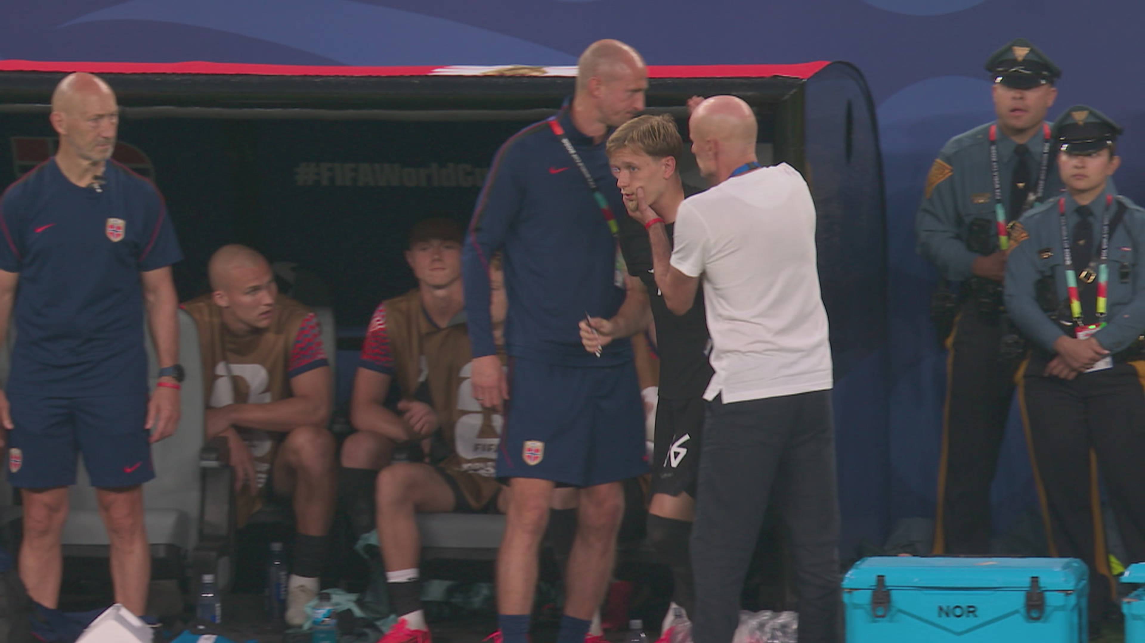 Ein fotballspelar blir tatt hand om av ein medarbeidar på benken. To personar står bak dei. Bakspillerne sit på benken. (Bildebeskrivelsen er laget av ein KI-teneste)