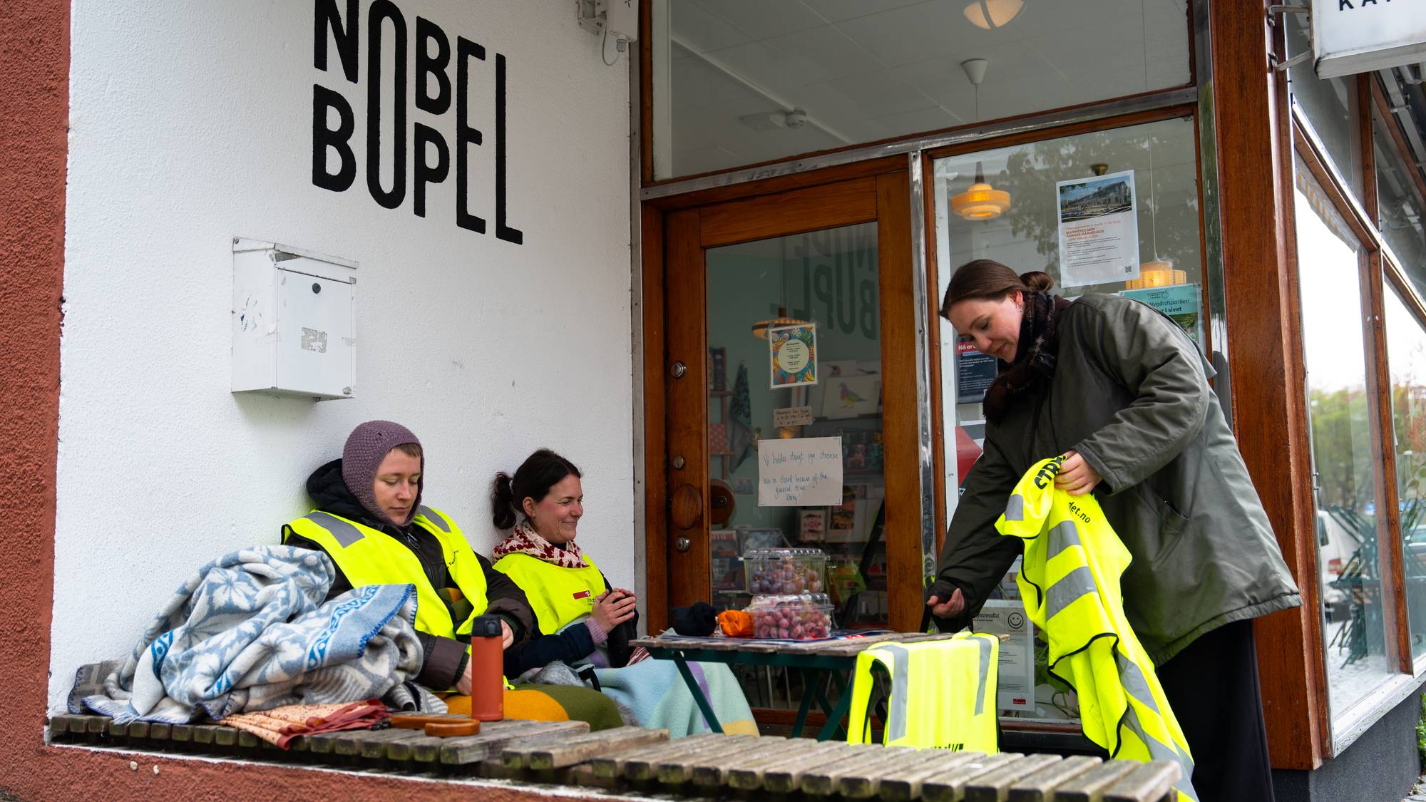 Tre personer sitter og står utenfor en kafé med skiltet "Nobel Bopel". To personer sitter på en benk med reflekser og tepper. En tredje person står og holder en gul refleksvest.