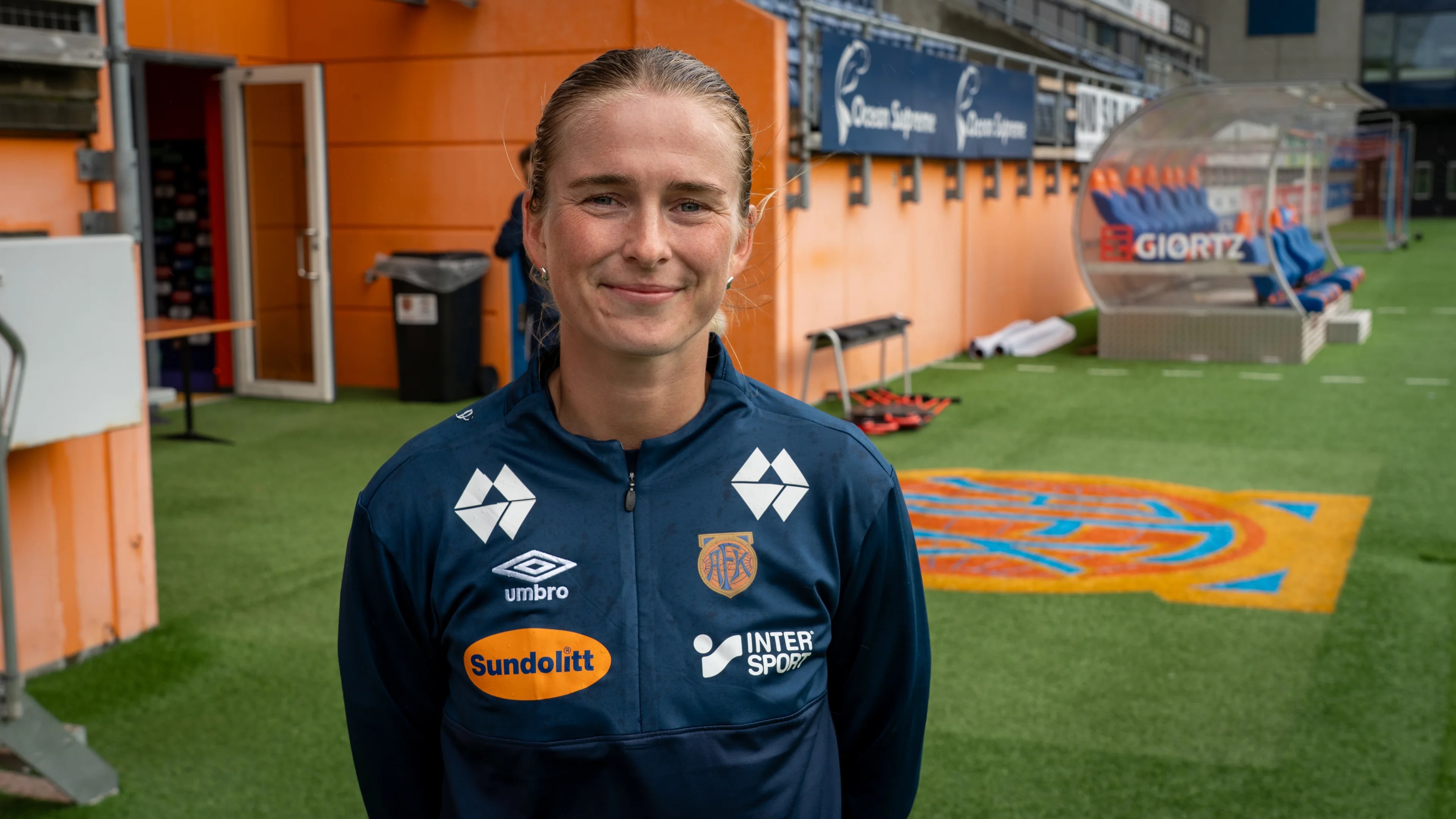 Fotballspiller i mørk blå treningsjakke med logoer på en fotballbane. (Bildebeskrivelsen er laget av en KI-tjeneste)