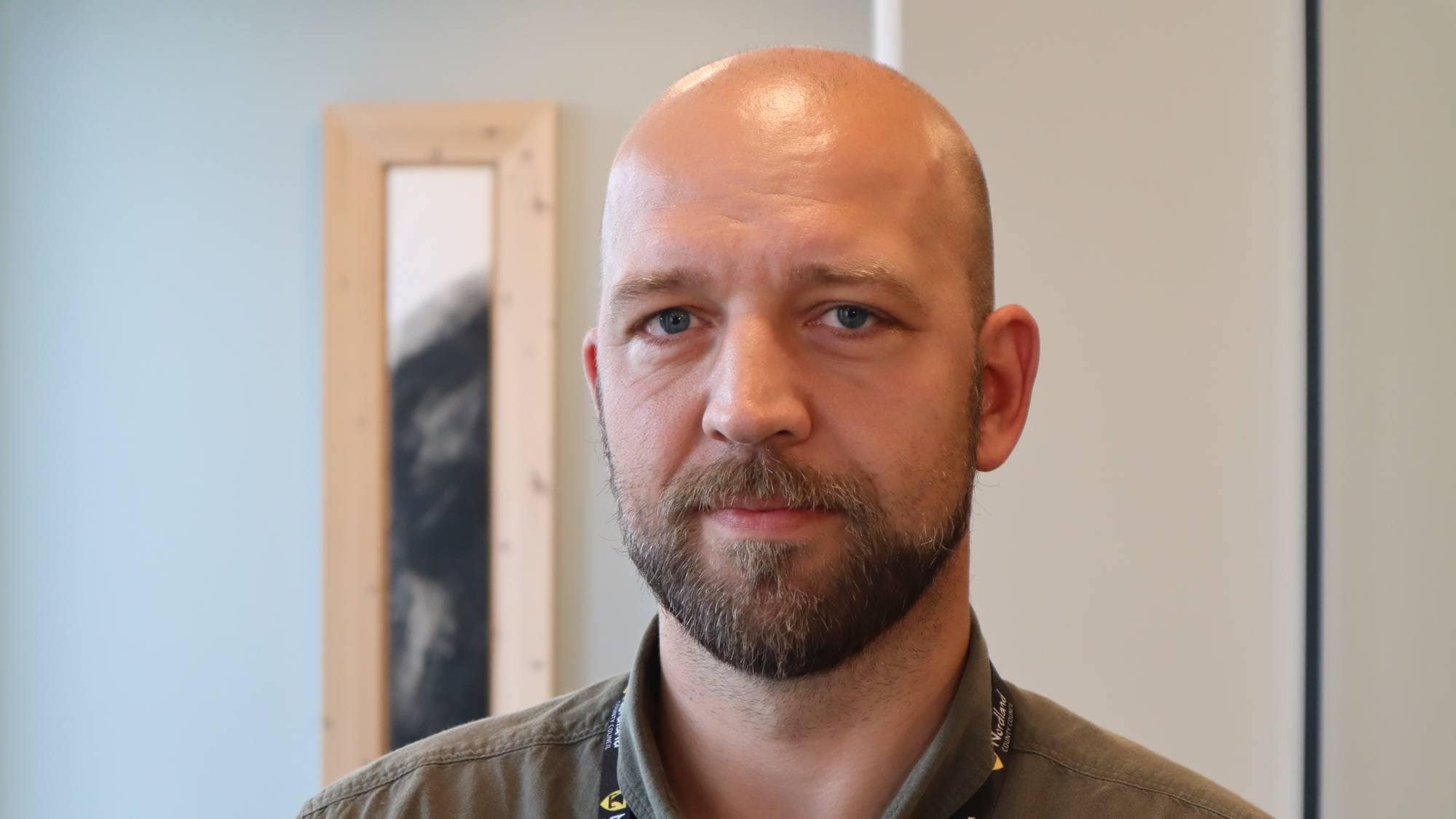 Portrett av en skalla mann med skjegg i en olivenfarget skjorte og nøkkelbånd fra Nordland fylkeskommune. (Bildebeskrivelsen er laget av en KI-tjeneste)