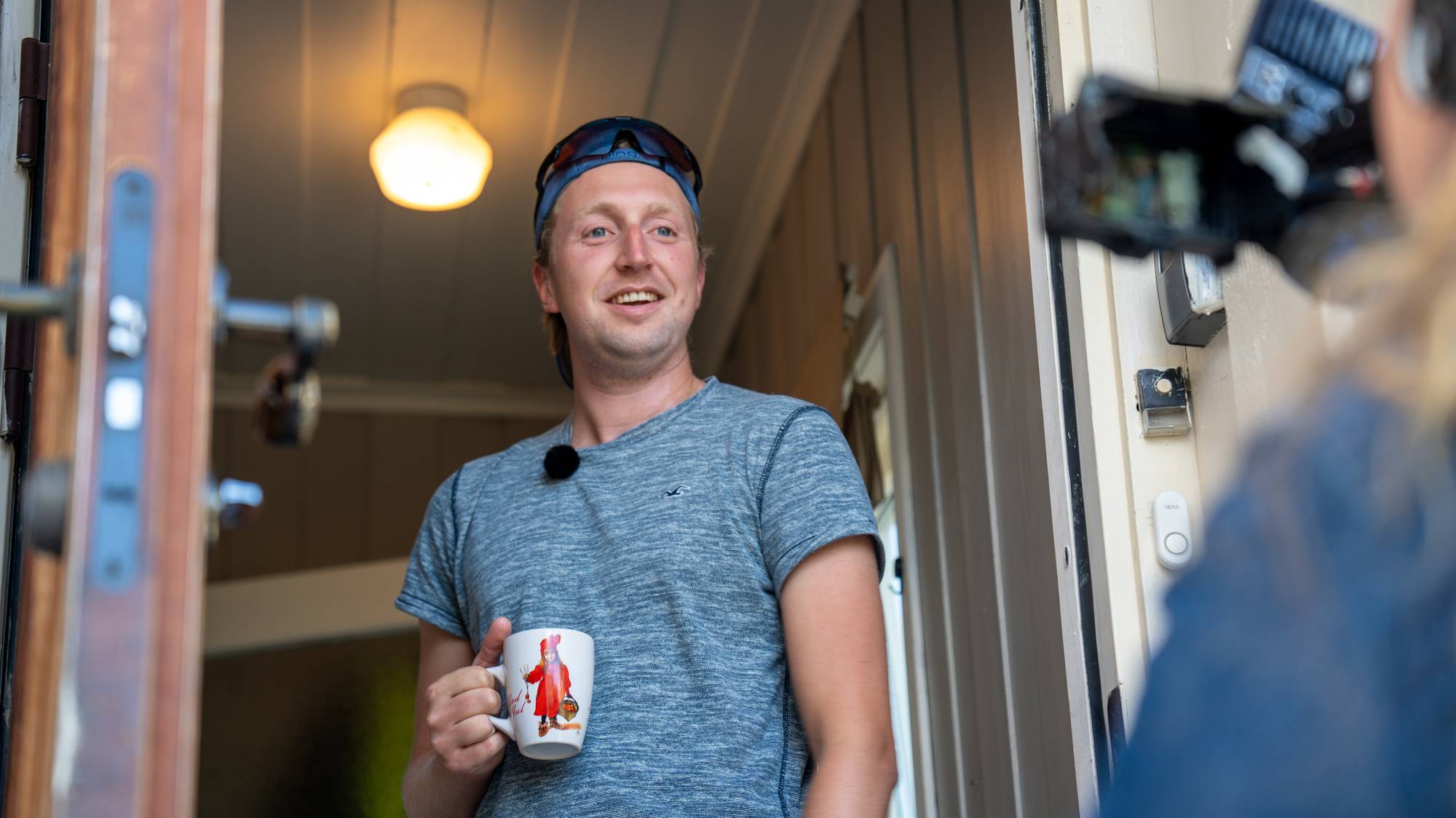 En mann med kaffekopp står i en døråpning mens han blir filmet. (Bildebeskrivelsen er laget av en KI-tjeneste)