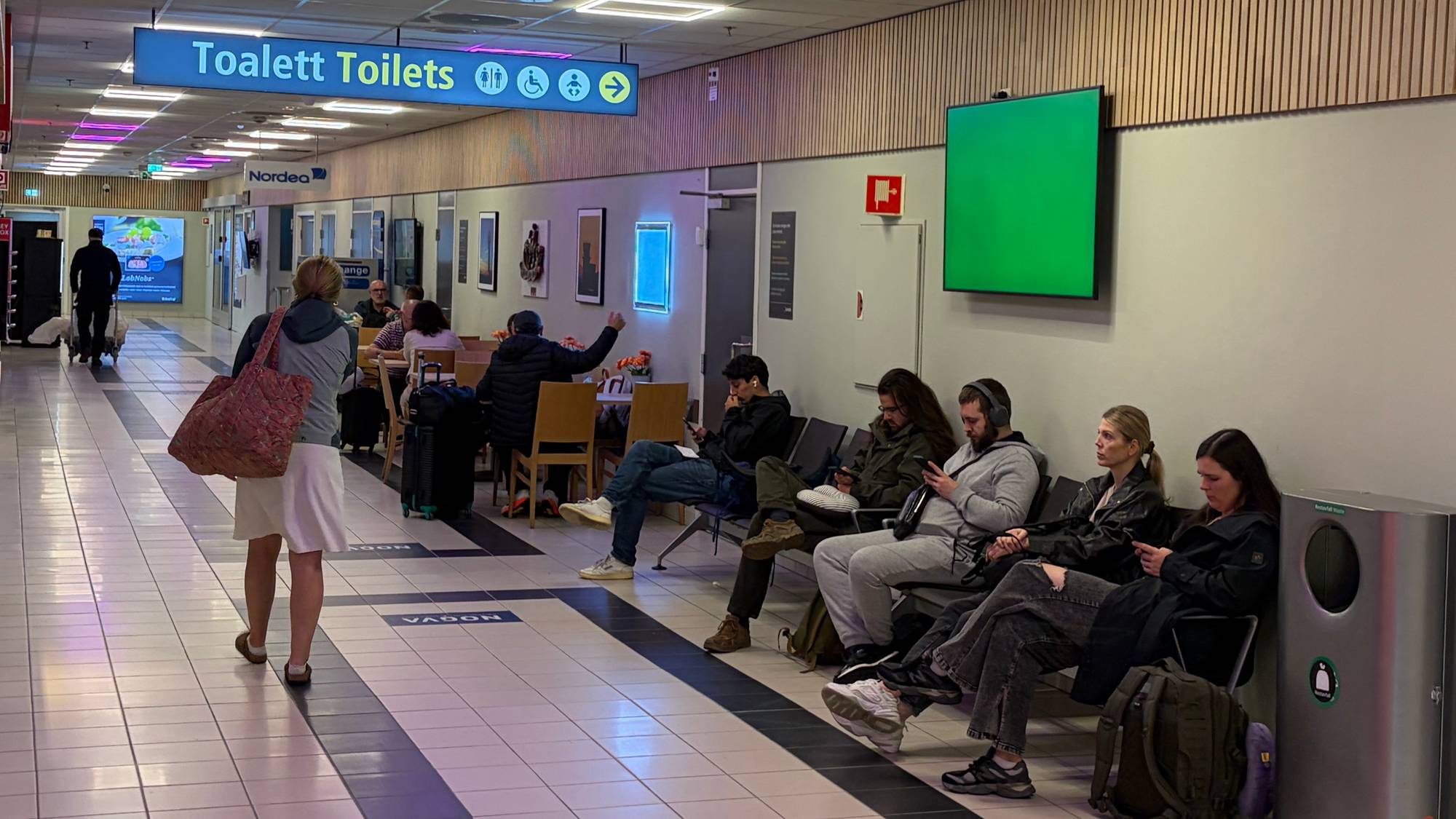 Personer venter på stoler i en terminalbygning. Skilt markerer vei til toaletter i korridoren. (Bildebeskrivelsen er laget av en KI-tjeneste)