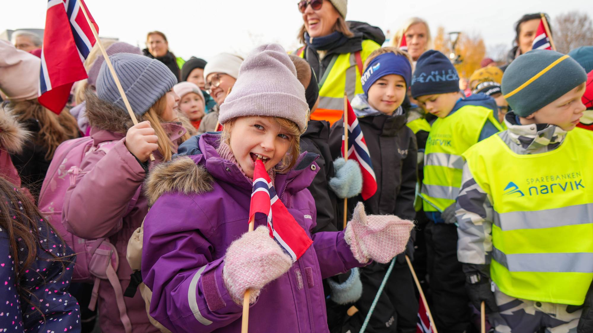 En jente med lue, votter og lilla vinterjakke holder et flagg blant flere andre barn med flagg.