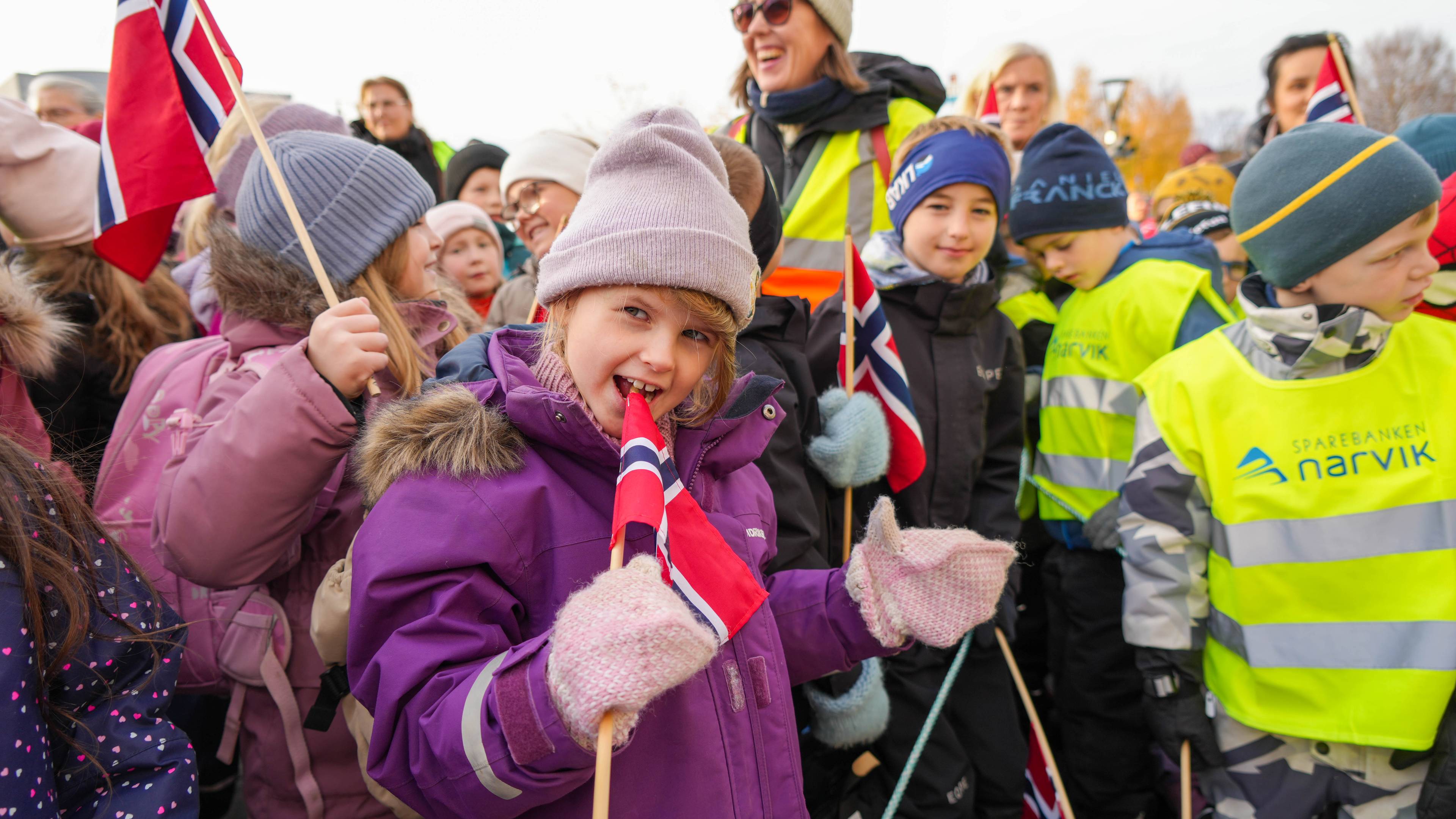 En jente med lue, votter og lilla vinterjakke holder et flagg blant flere andre barn med flagg.