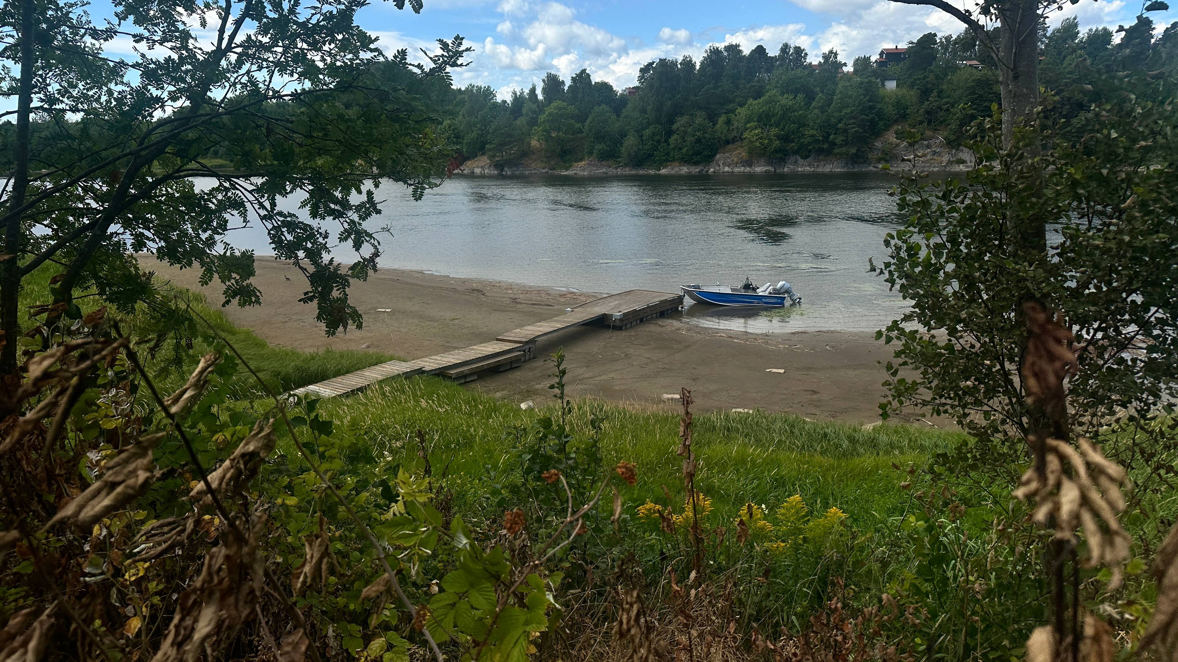 En liten båt ligger fortøyd ved en trebrygge på en sandstrand ved en elv omgitt av vegetasjon. (Bildebeskrivelsen er laget av en KI-tjeneste)