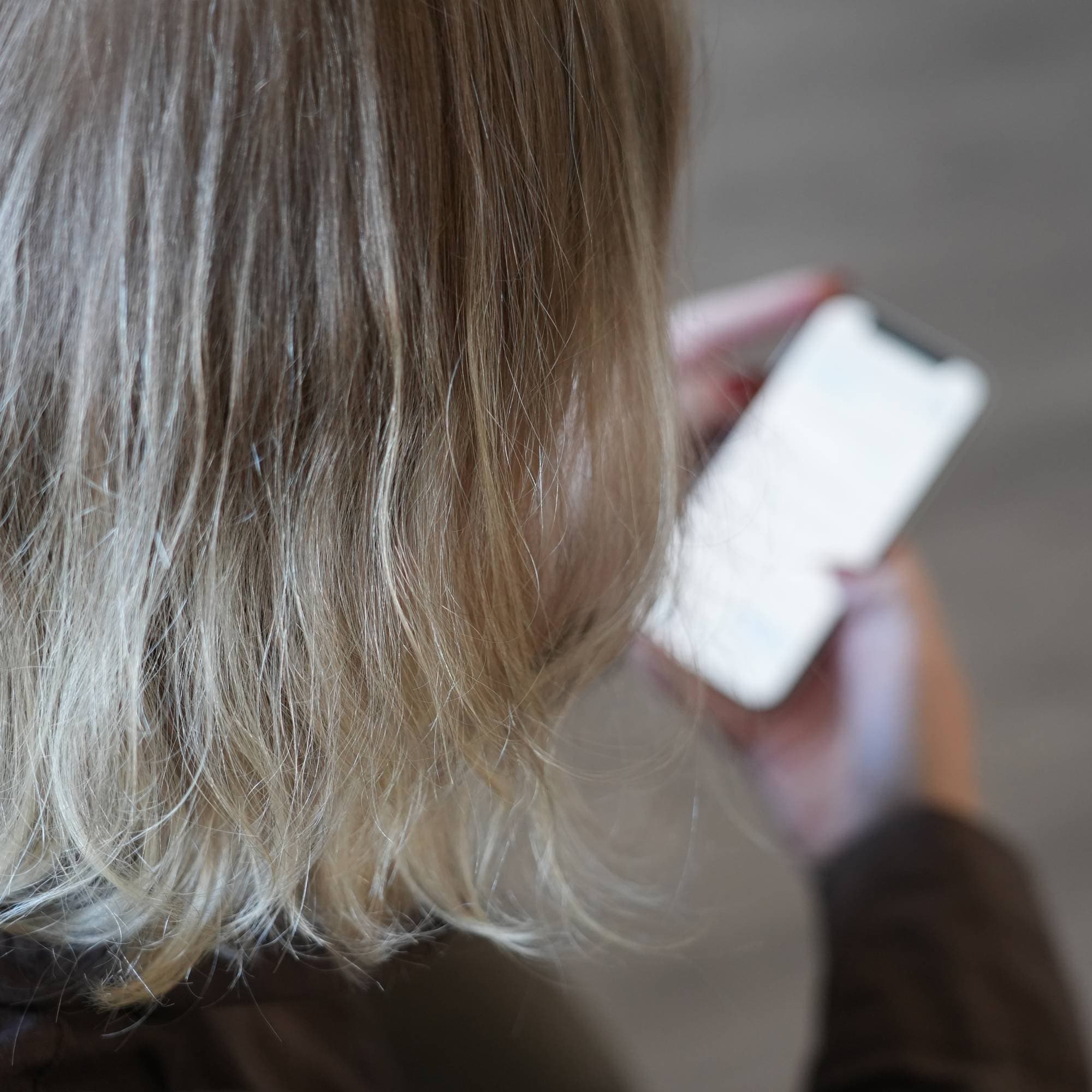 En uklar mobilskjerm vises over skulderen på en kvinne. Fokus i bildet er det blonde håret