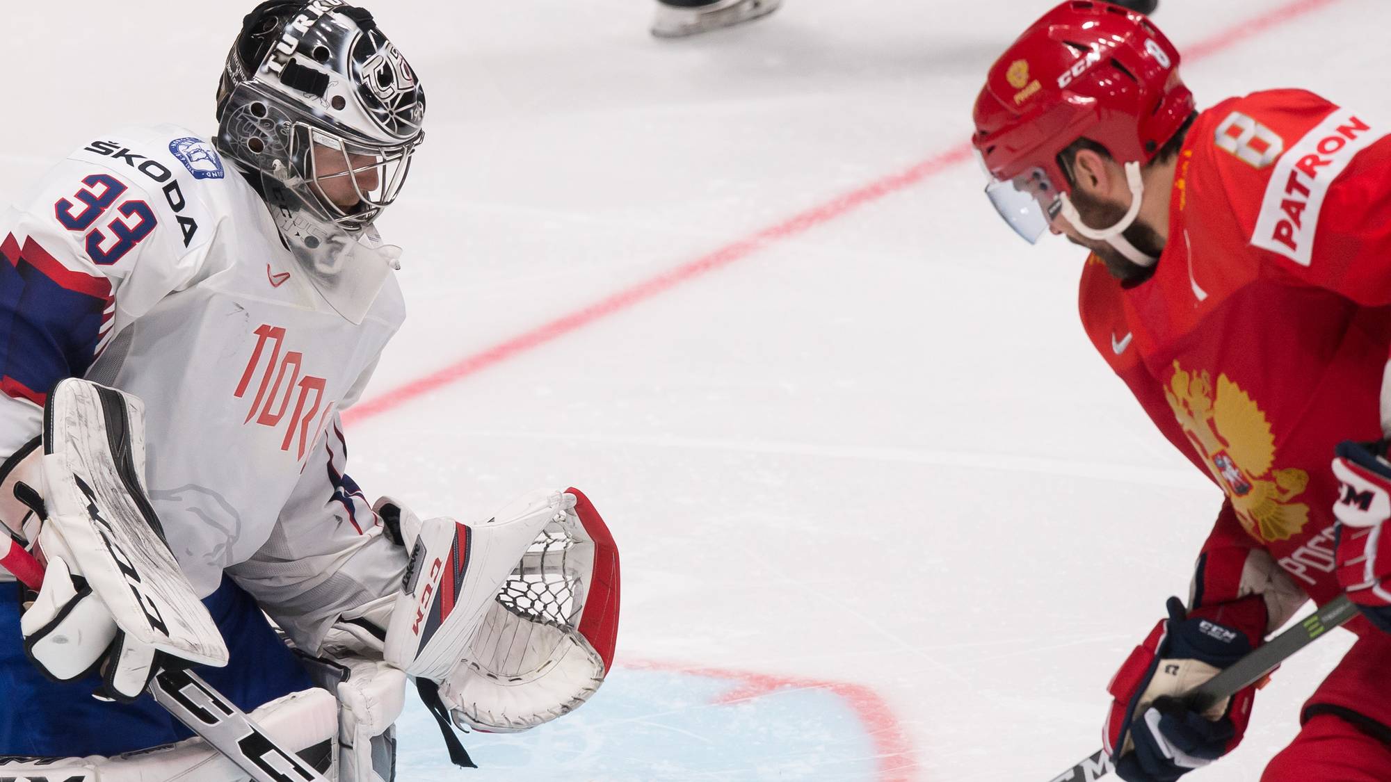 En ishockeymålvakt i hvit drakt med nummer 33 og en russisk spiller i rød drakt under en kamp. Spilleren skyter pucken mot målet der målvakten forsvarer. Bildet er tatt på isen i en hockeyhall. (Bildebeskrivelsen er laget av en KI-tjeneste)