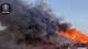Foto: Politiet En stor rød låve brenner og store mengder mørk røyk stiger opp mot en klar himmel. Flammene slikker taket på bygningen. Foran låven står flere store tanker. Et politiemblem er synlig øverst til venstre. (Bildebeskrivelsen er laget av en KI-tjeneste)