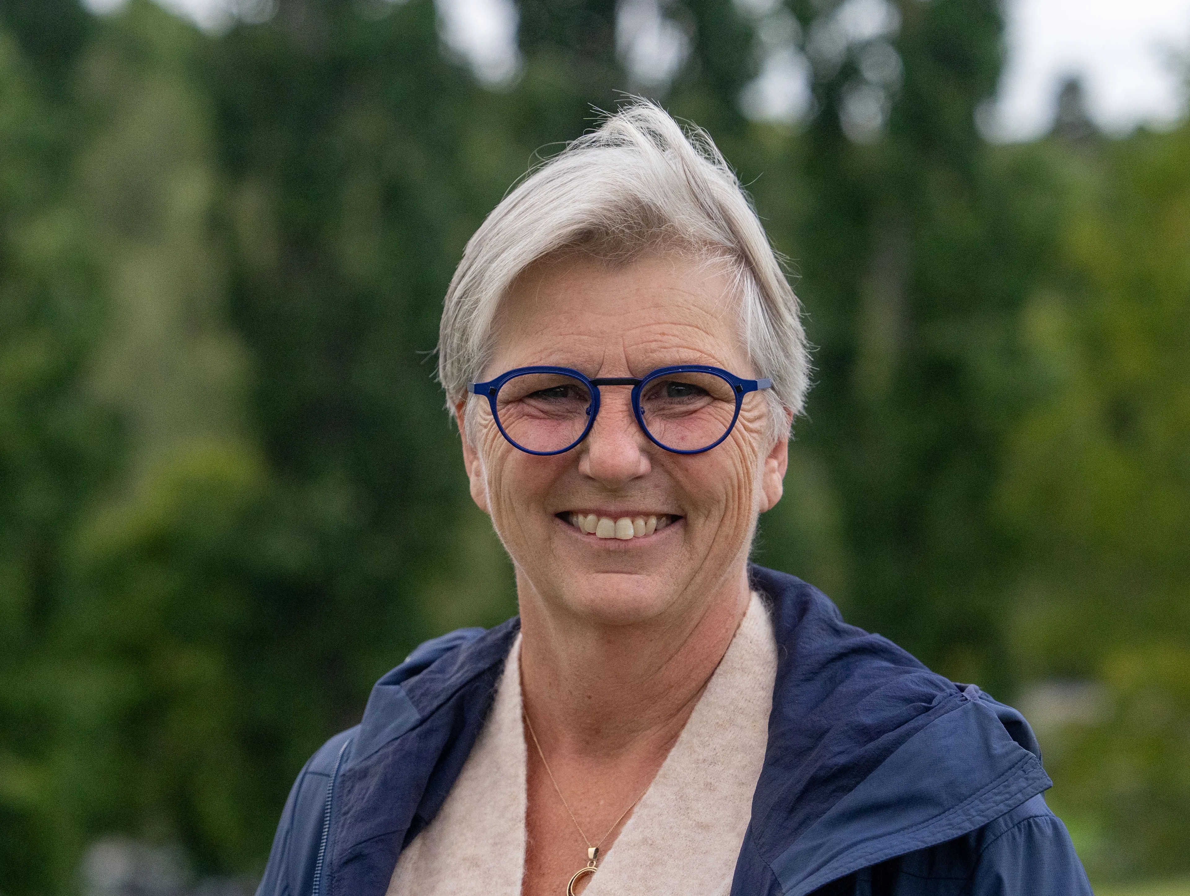 En kvinne med kort lyst hår, blå briller og blå jakke står ute foran grønne trær og smiler mot kameraet.